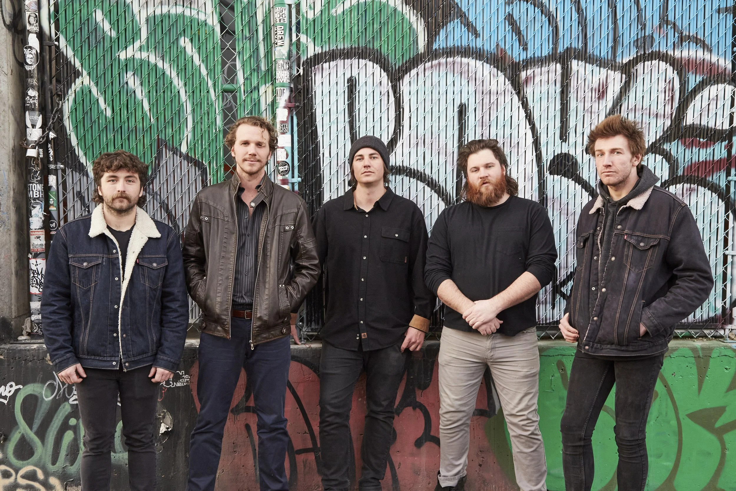 Five men standing in front of a graffiti-covered metal fence, dressed in casual clothing including jackets and hoodies.