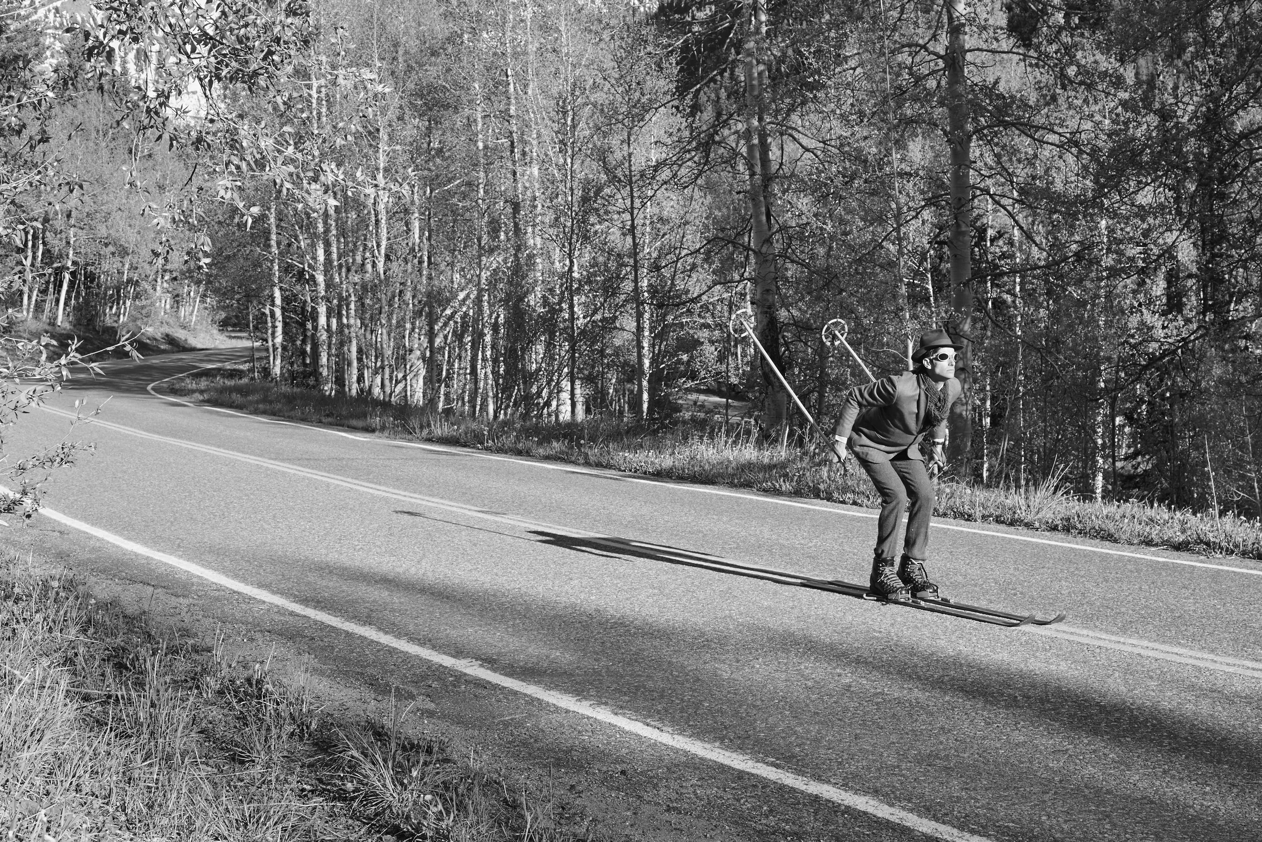 A person wearing a hat, sunglasses, and roller skis is skating on a curved mountain road surrounded by trees.