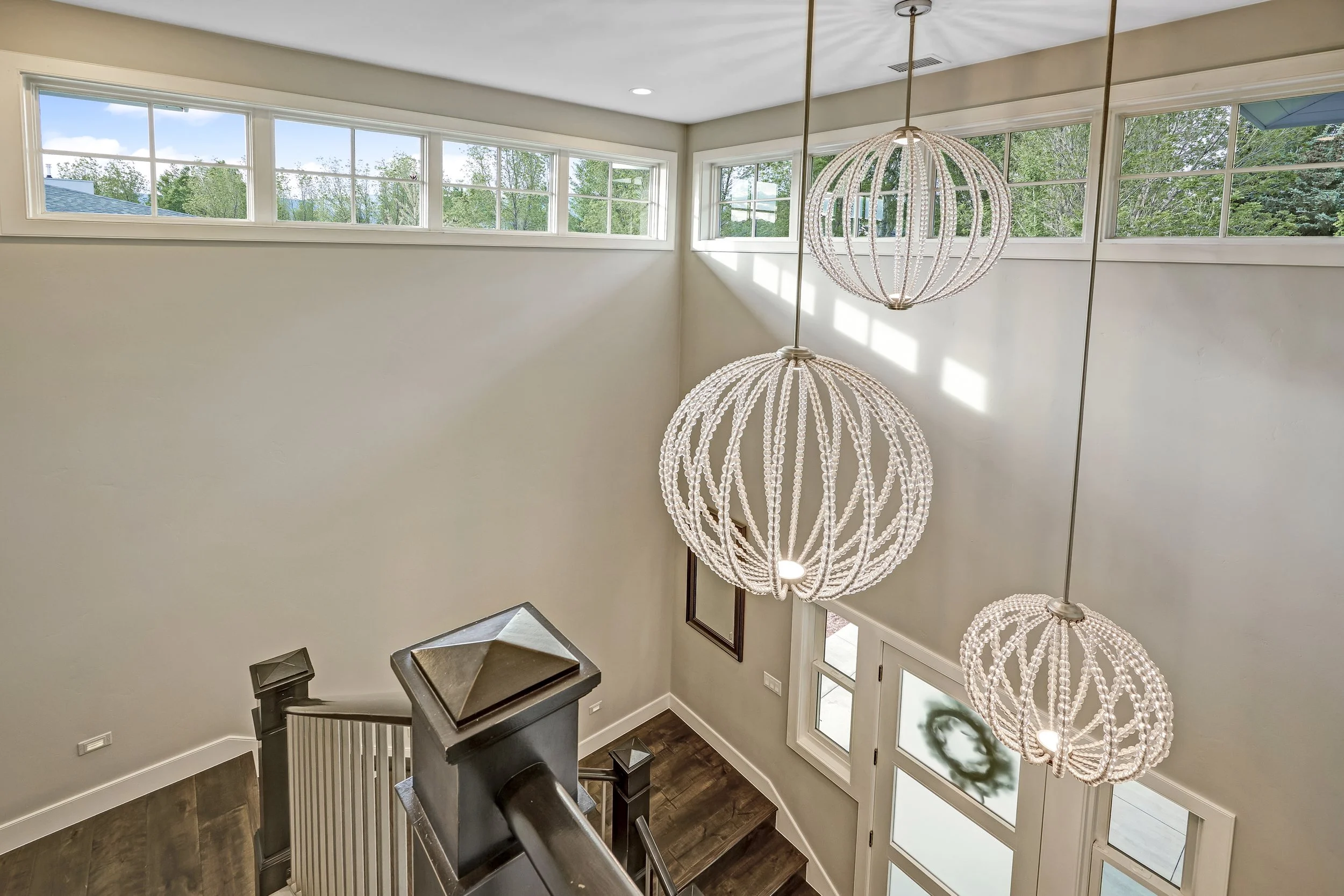 Interior of a house showing a staircase with a large spherical chandelier hanging above, high windows, and a staircase landing with dark wood flooring.