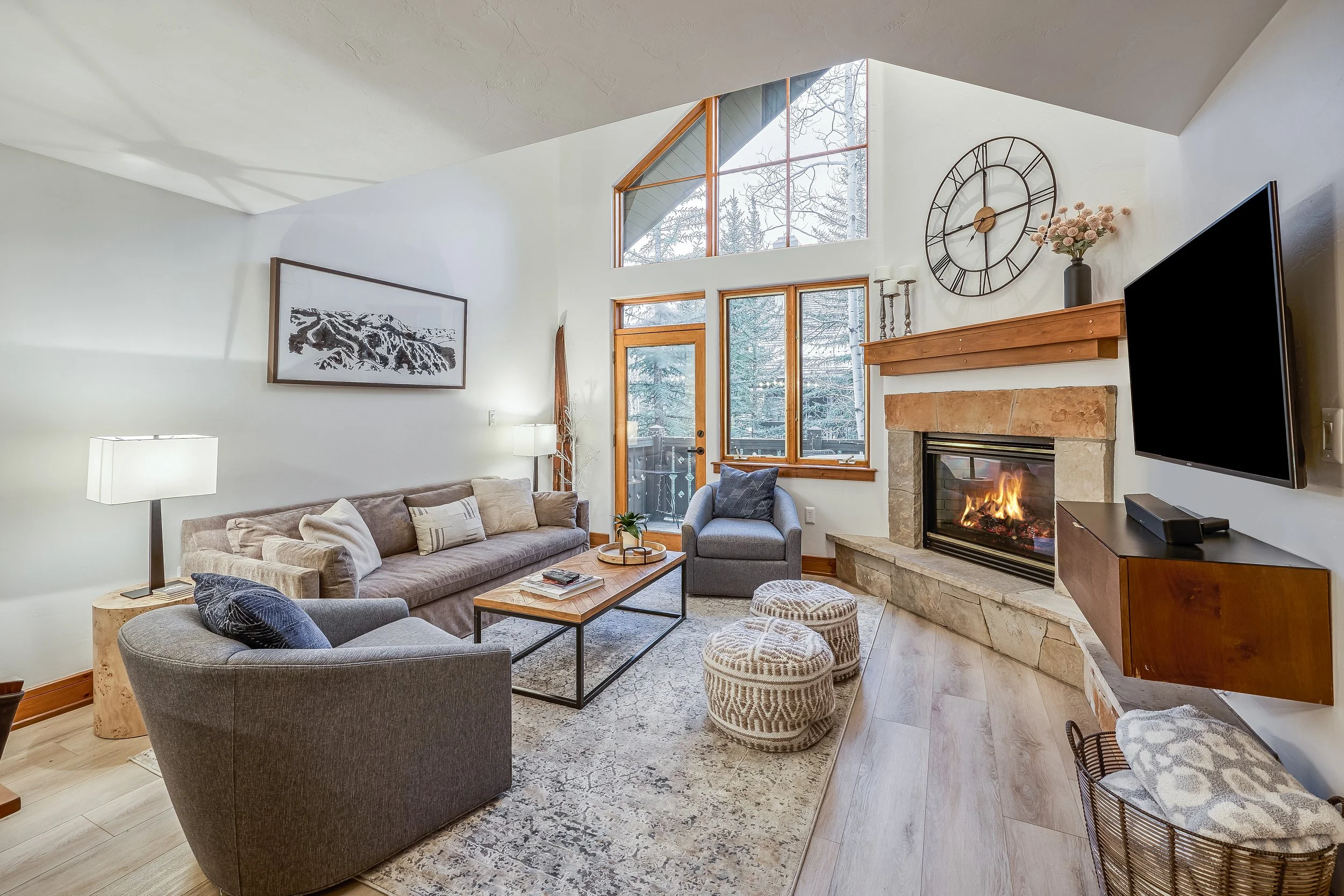 Living room with high vaulted ceiling, large windows, and a stone fireplace with a fire burning. Contains a gray armchair, a beige sofa with pillows, a coffee table with magazines, two patterned poufs, and a side table with a lamp. Wall decor include