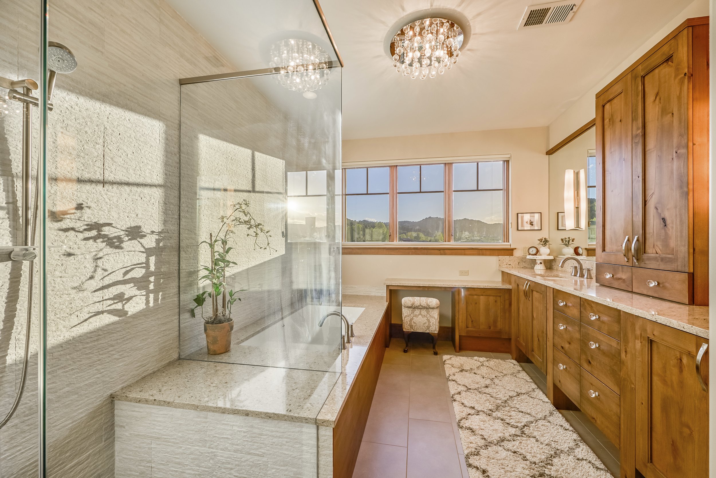 Bright bathroom with large window and mountain view, featuring a glass-enclosed shower with white stone walls, wooden cabinetry, and chandelier lighting.