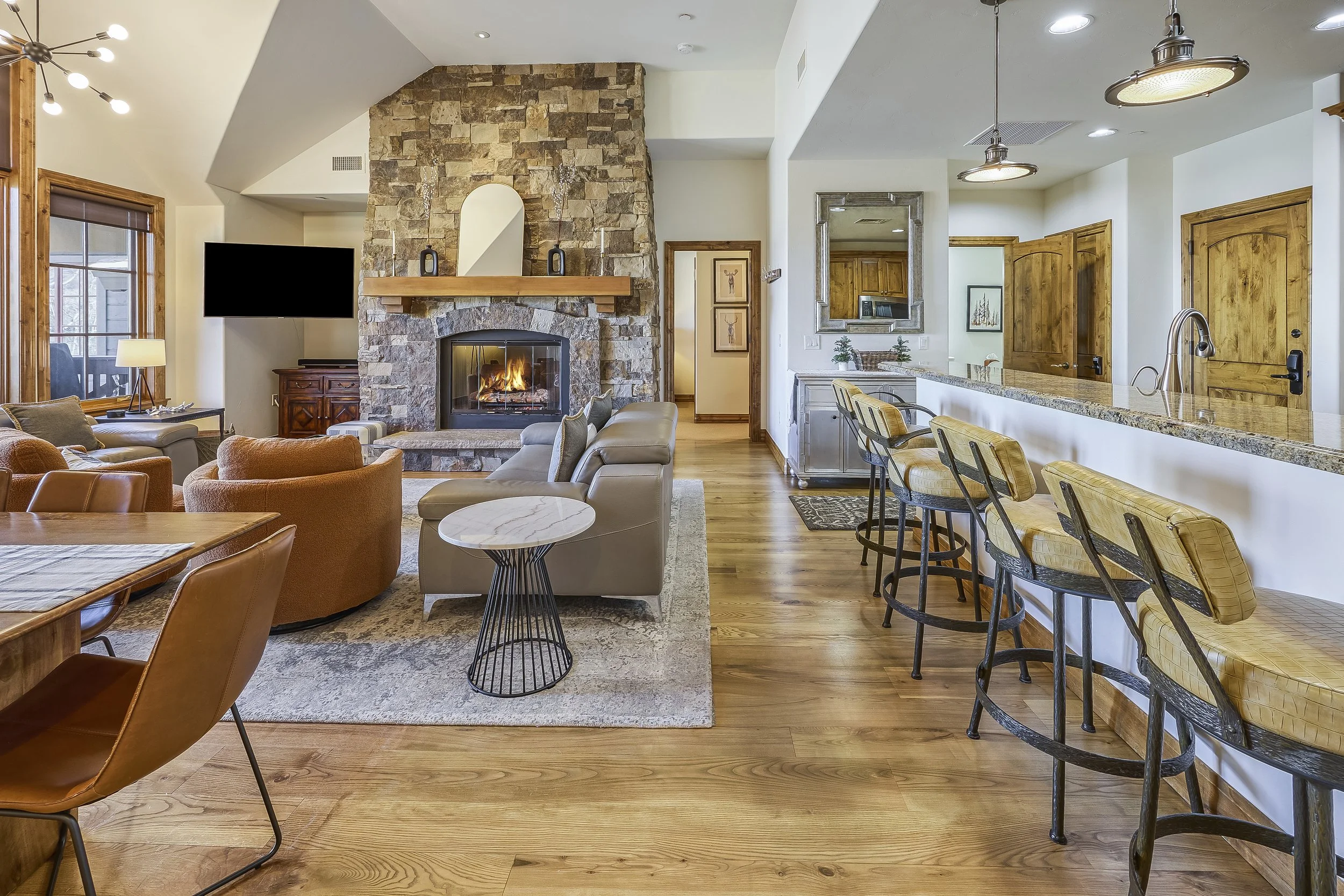 A living room with a stone fireplace, a mounted flat-screen TV, a seating area with armchairs and a sofa, a marble-topped coffee table, and a view into a kitchen with bar stools at a granite countertop.