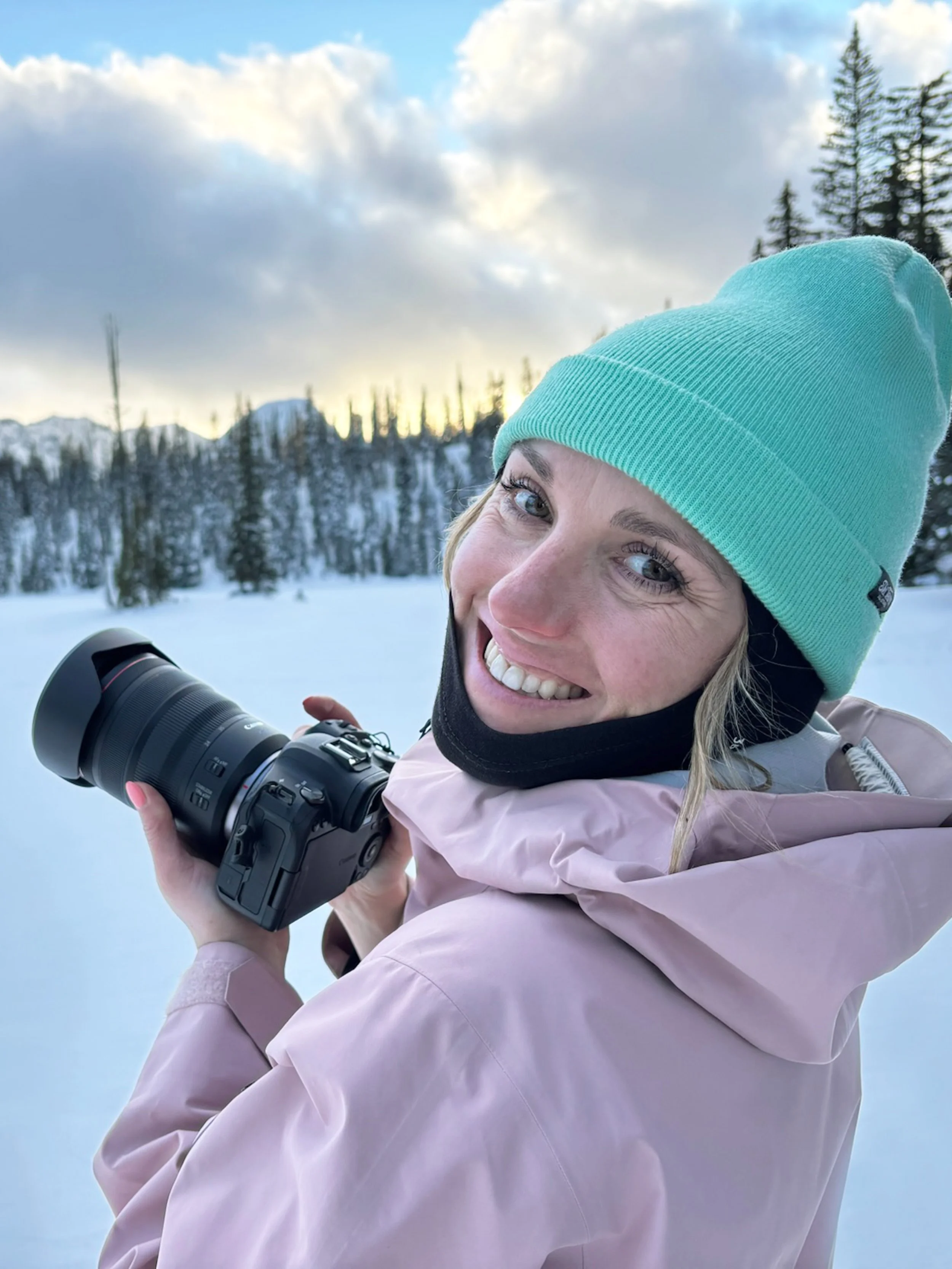 A woman smiling in winter clothing, holding a camera outdoors in a snowy landscape with pine trees and mountains in the background.