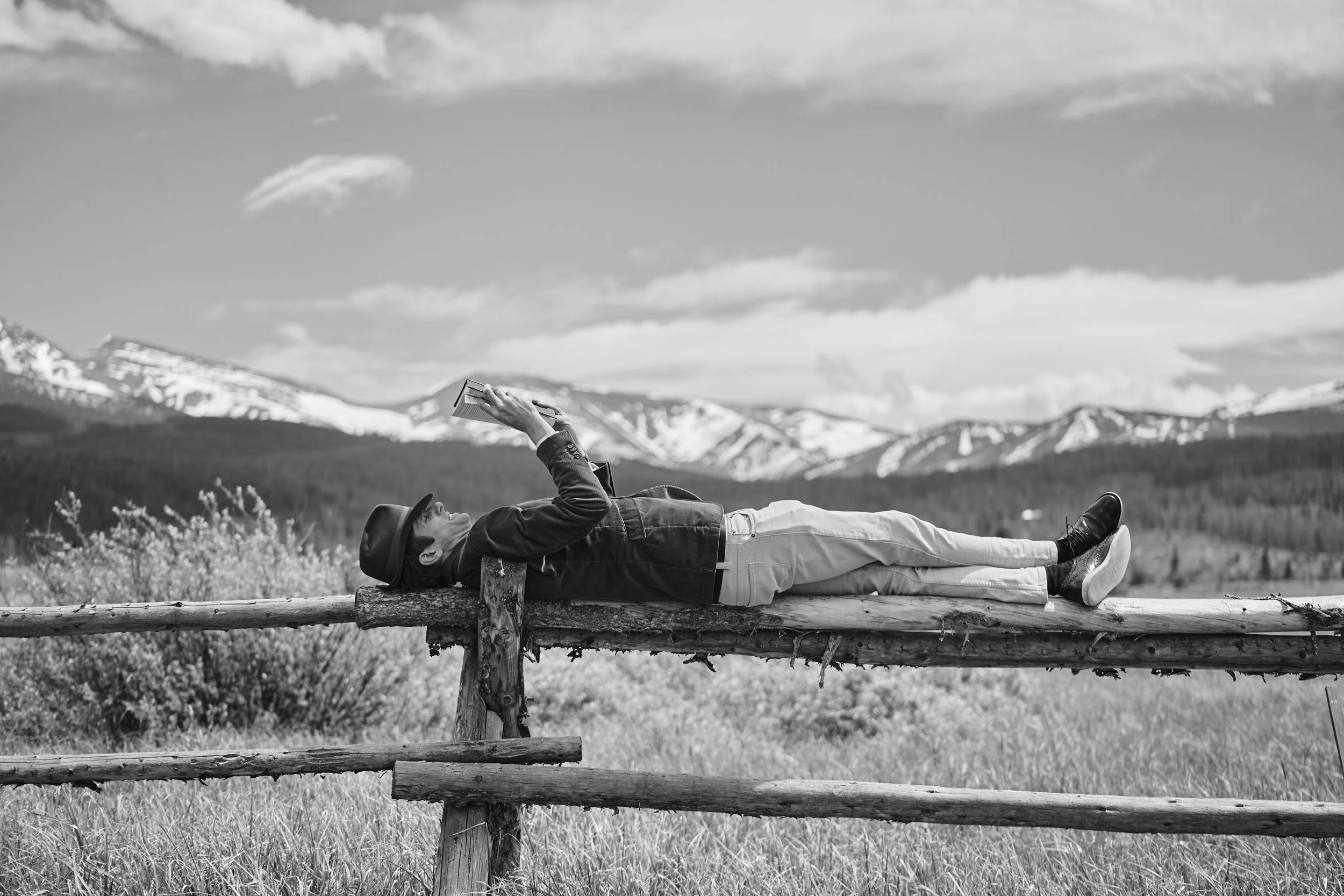 A person lying on a wooden fence in a grassy field, reading a book, with mountains and a partly cloudy sky in the background.