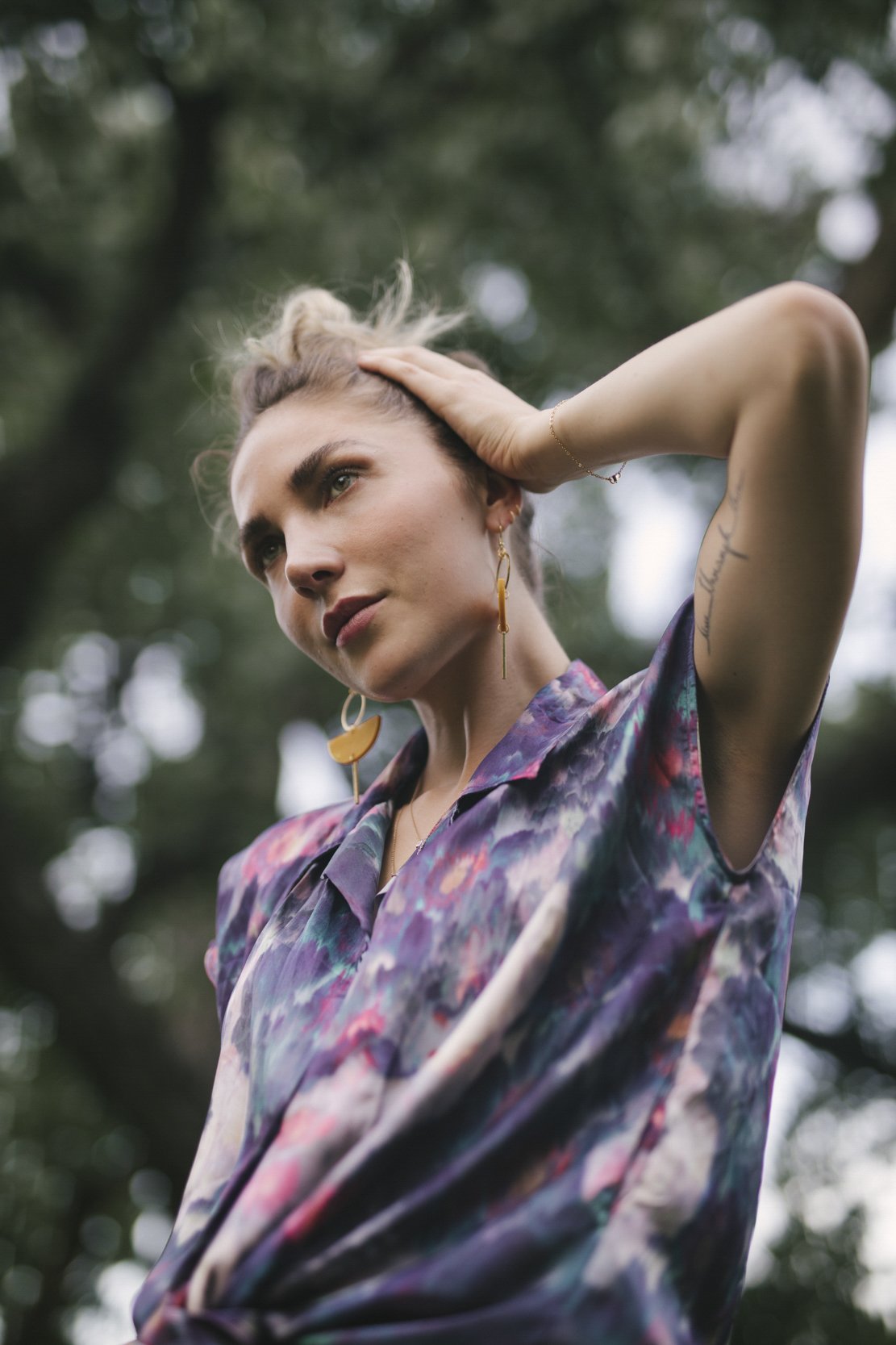A young woman with blonde hair styled in an updo, wearing colorful earrings and a multicolored floral-patterned blouse, posing outdoors with trees in the background.
