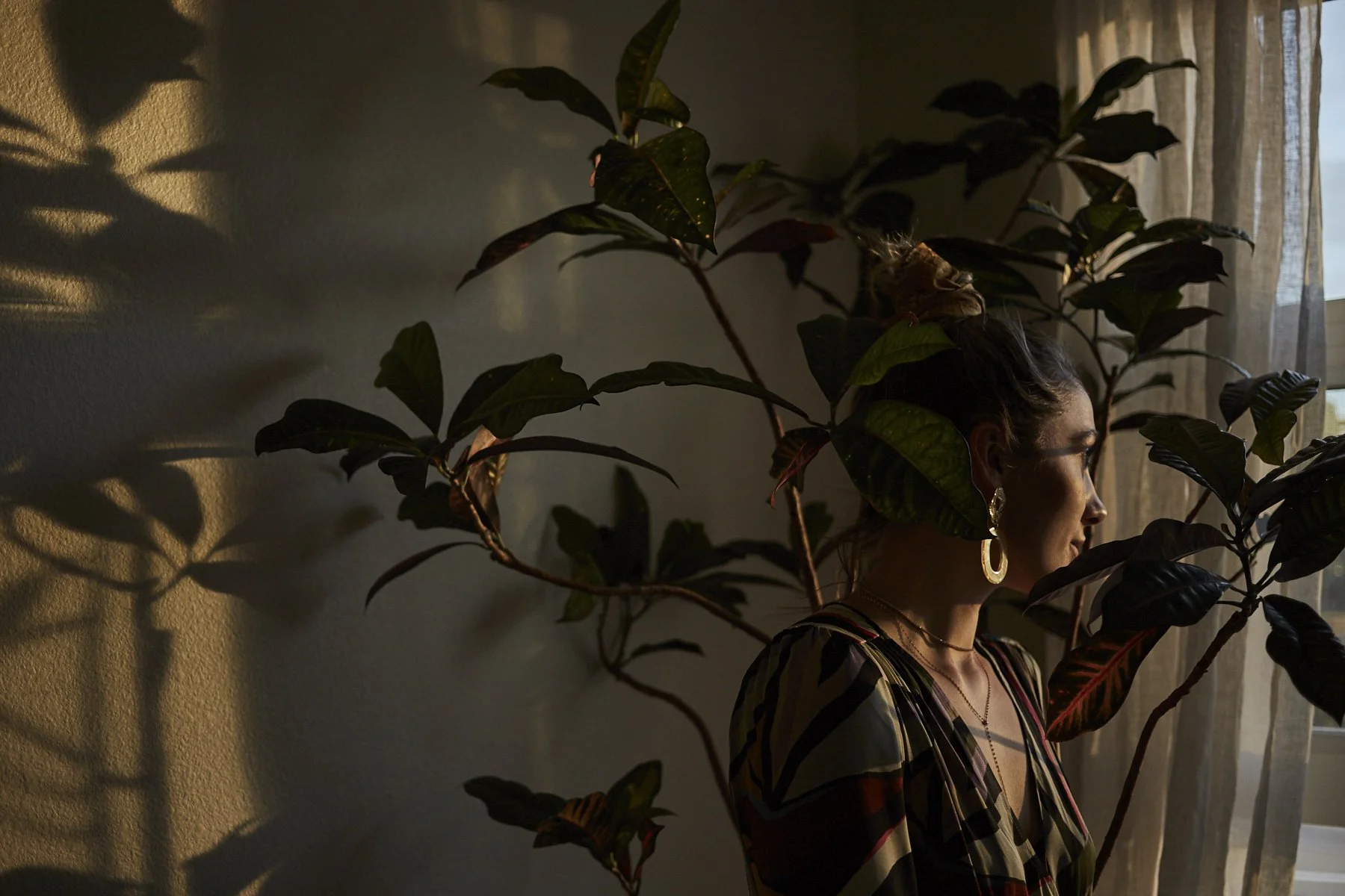 A woman with earrings and a necklace stands indoors by a window, partially obscured behind a large leafy plant, with sunlight casting shadows on the wall.