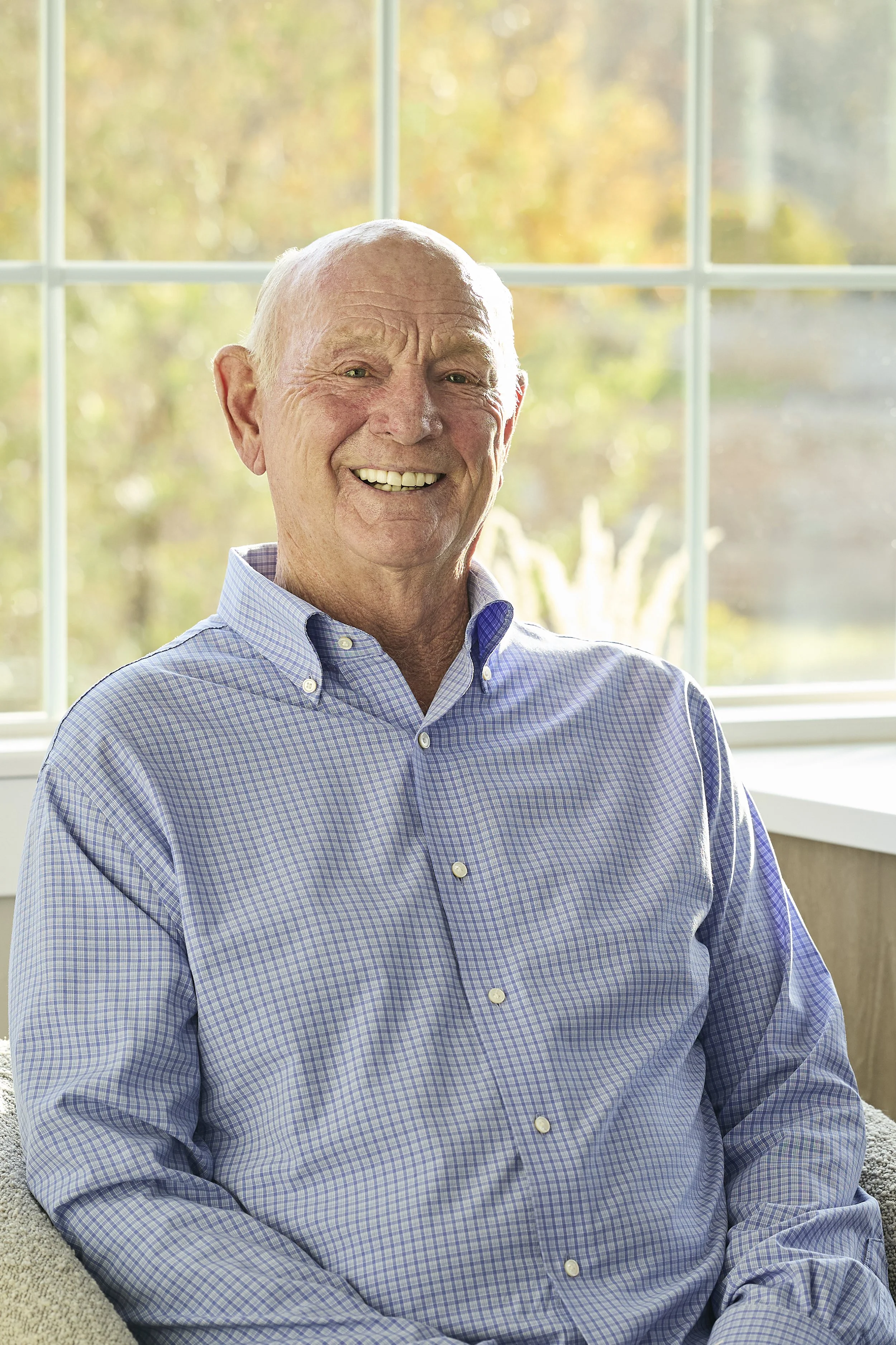An elderly man with a big smile, wearing a blue checkered button-up shirt, sitting indoors near large windows with a blurred background of trees and sunlight.