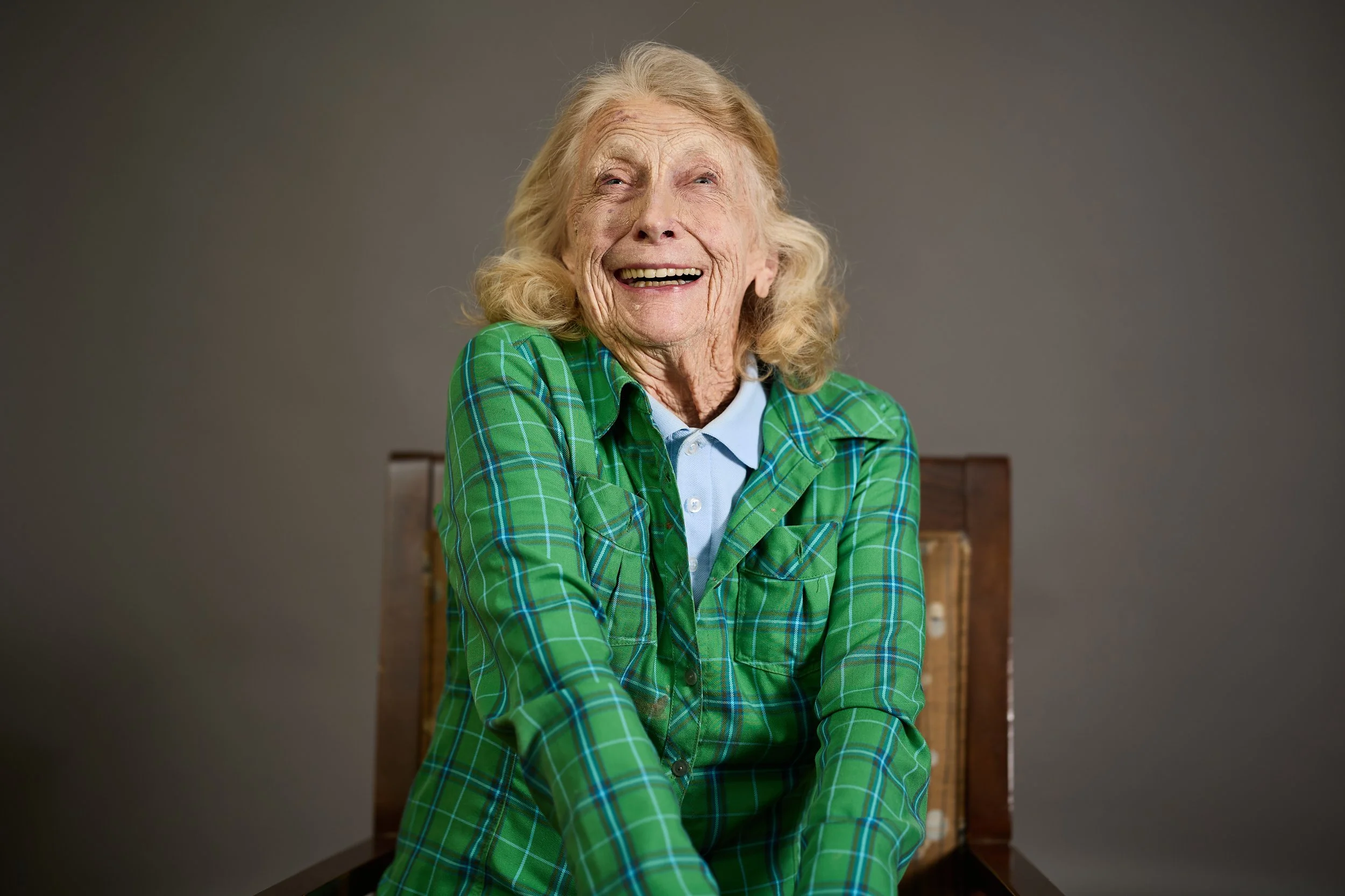 An elderly woman with long curly blonde hair smiling and looking upwards. She is wearing a green plaid shirt over a light blue collared shirt and sitting at a wooden table against a plain gray background.