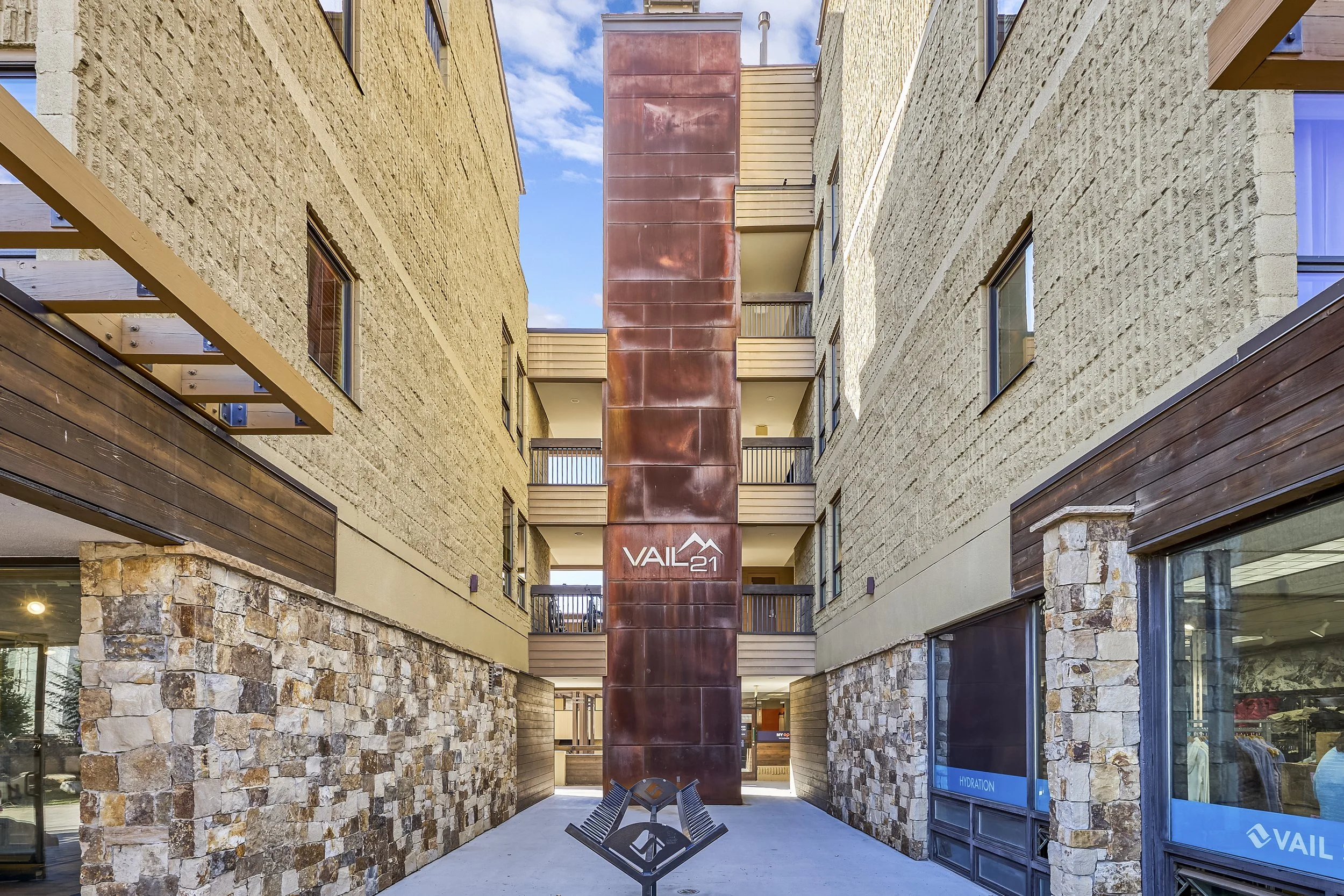 View of a modern multi-story apartment building with stone and brick facade. The building has balconies and a central rust-colored vertical panel with the Vail logo at the bottom. The sky is partly cloudy with patches of blue visible.