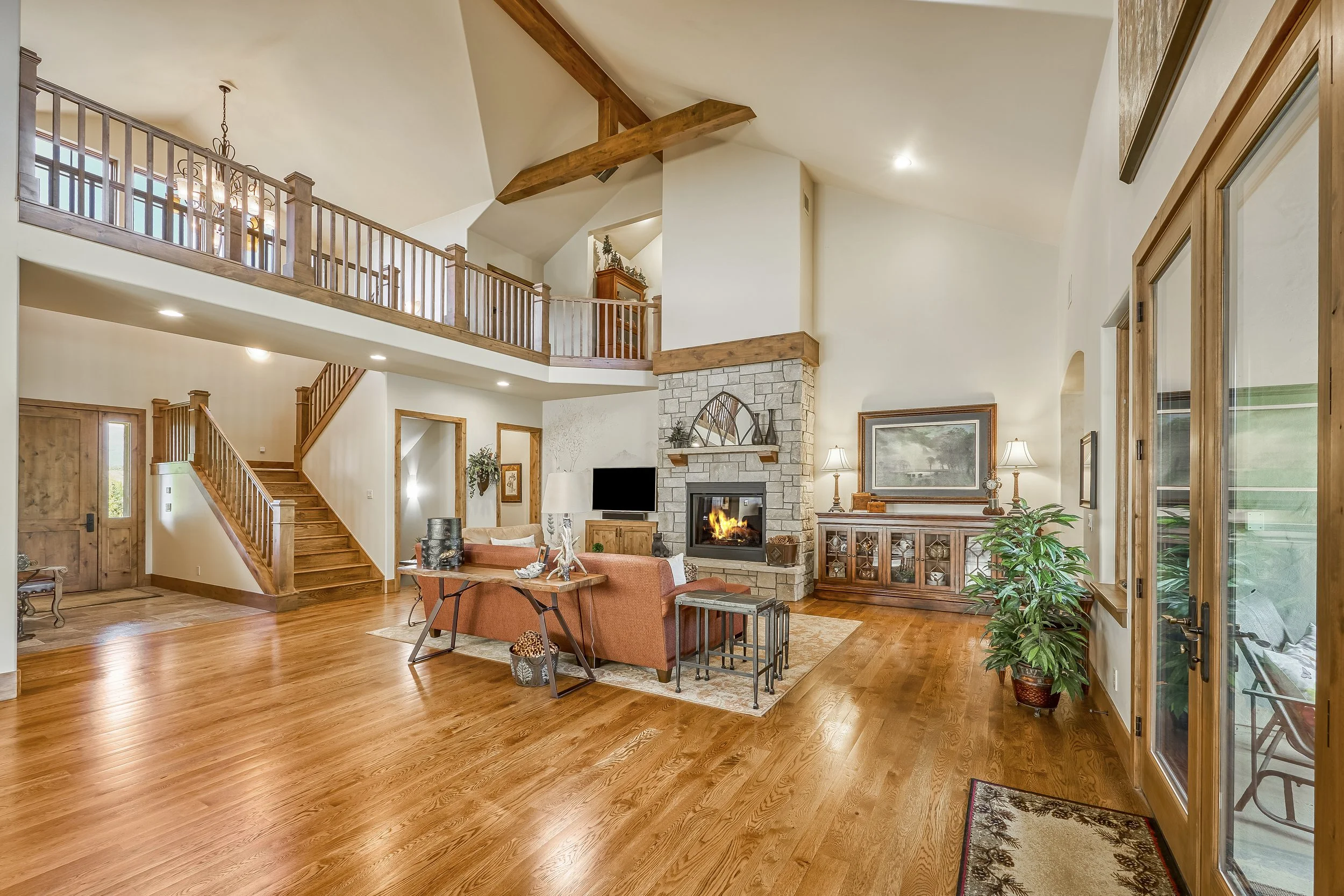 Interior of a spacious living room with a high ceiling, wooden floors, a stone fireplace, and wooden beams. There is furniture including a sofa and chairs, a wooden sideboard, and a glass door leading outside. A staircase with wooden railings is visi