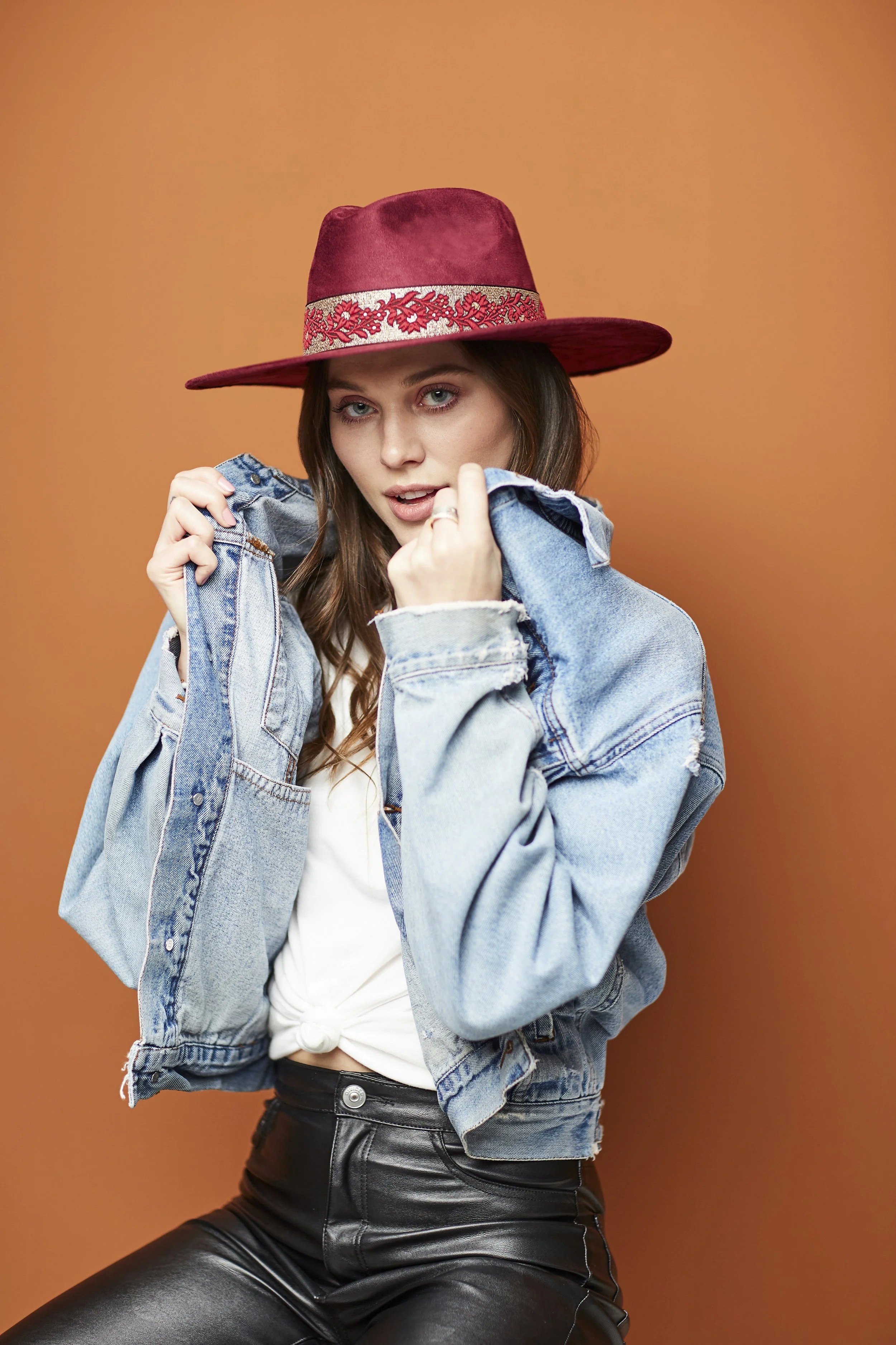 A young woman wearing a red wide-brimmed hat with decorative band, a light denim jacket, and black leather pants, standing against an orange background.
