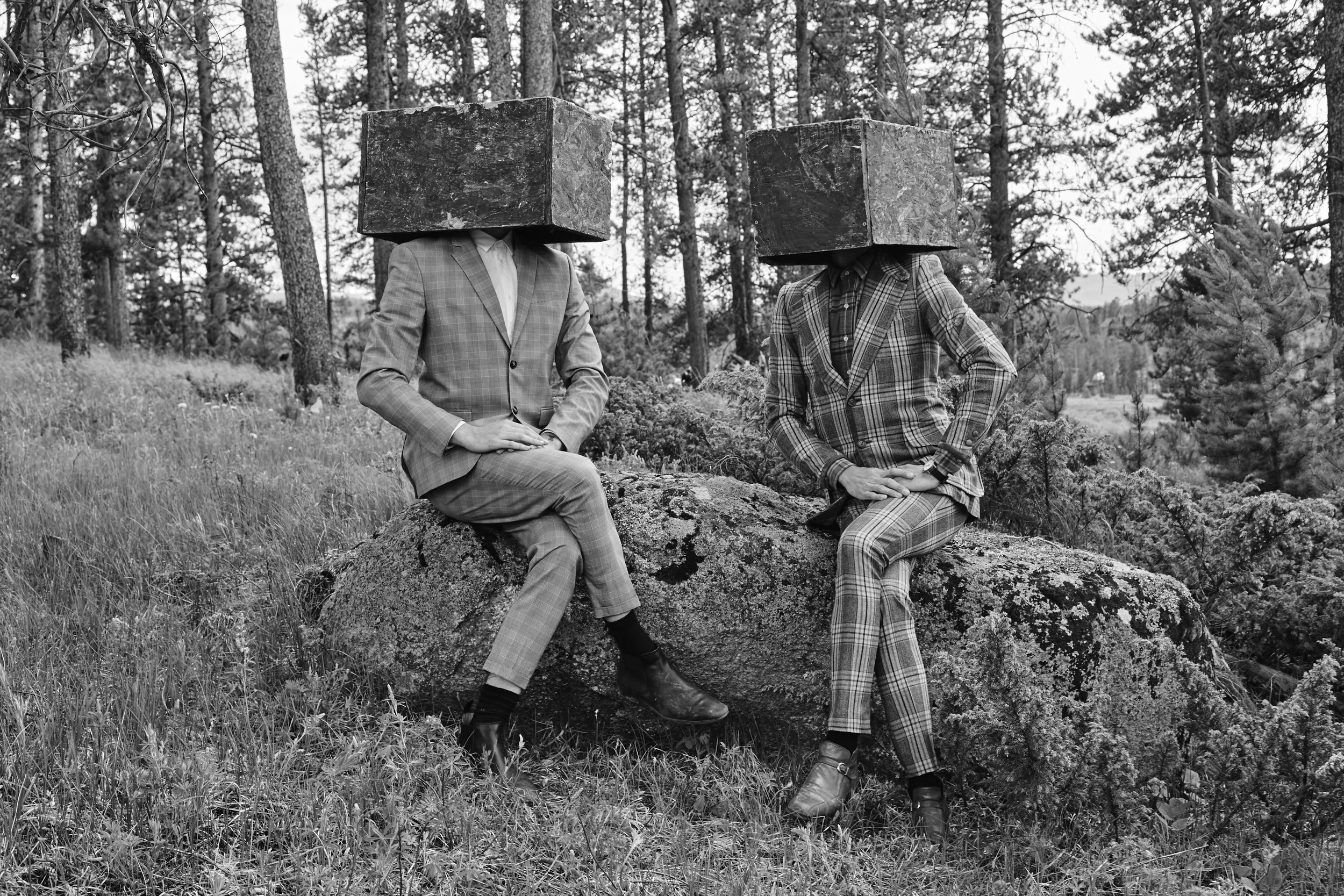 Two people wearing suits with large cube boxes on their heads, sitting on a rock in a forest.
