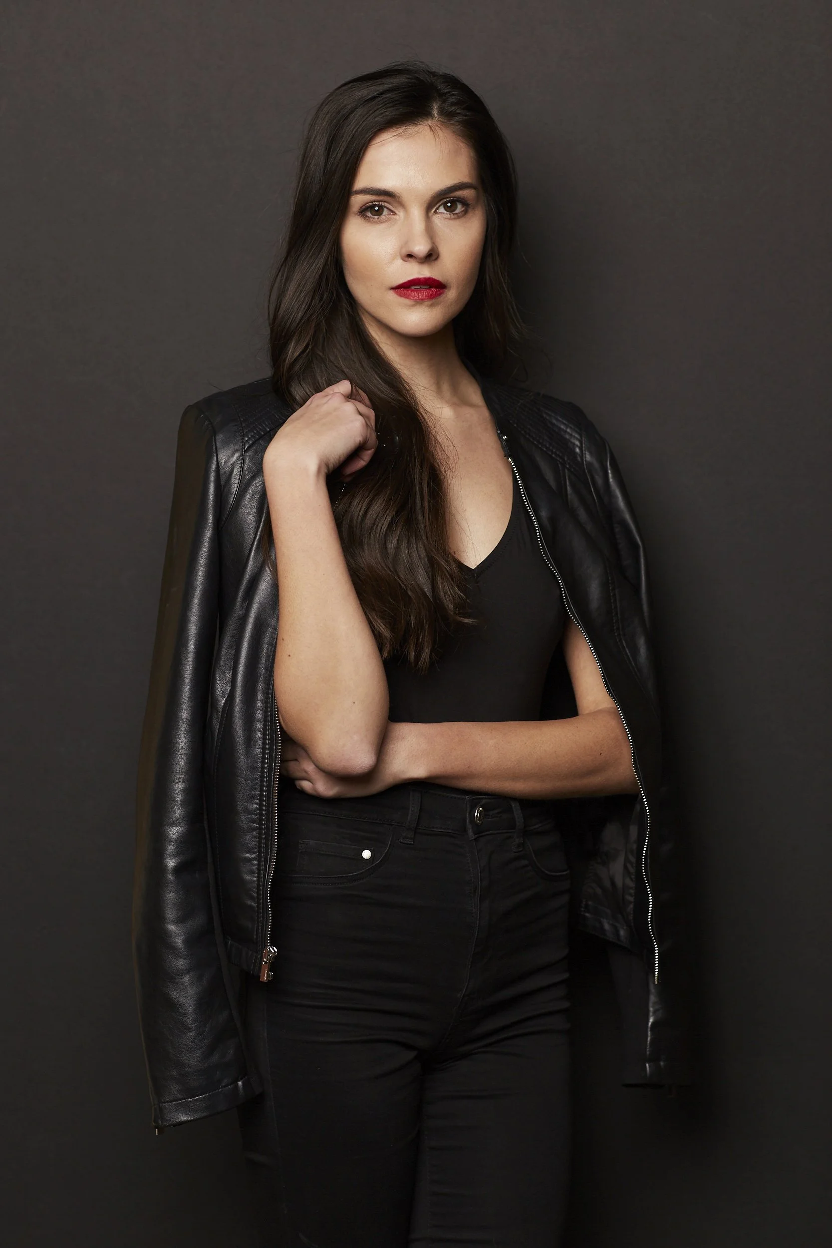 A woman with long dark hair wearing a black leather jacket over a black top stands in front of a dark background. She has her hand near her shoulder and is looking directly at the camera.