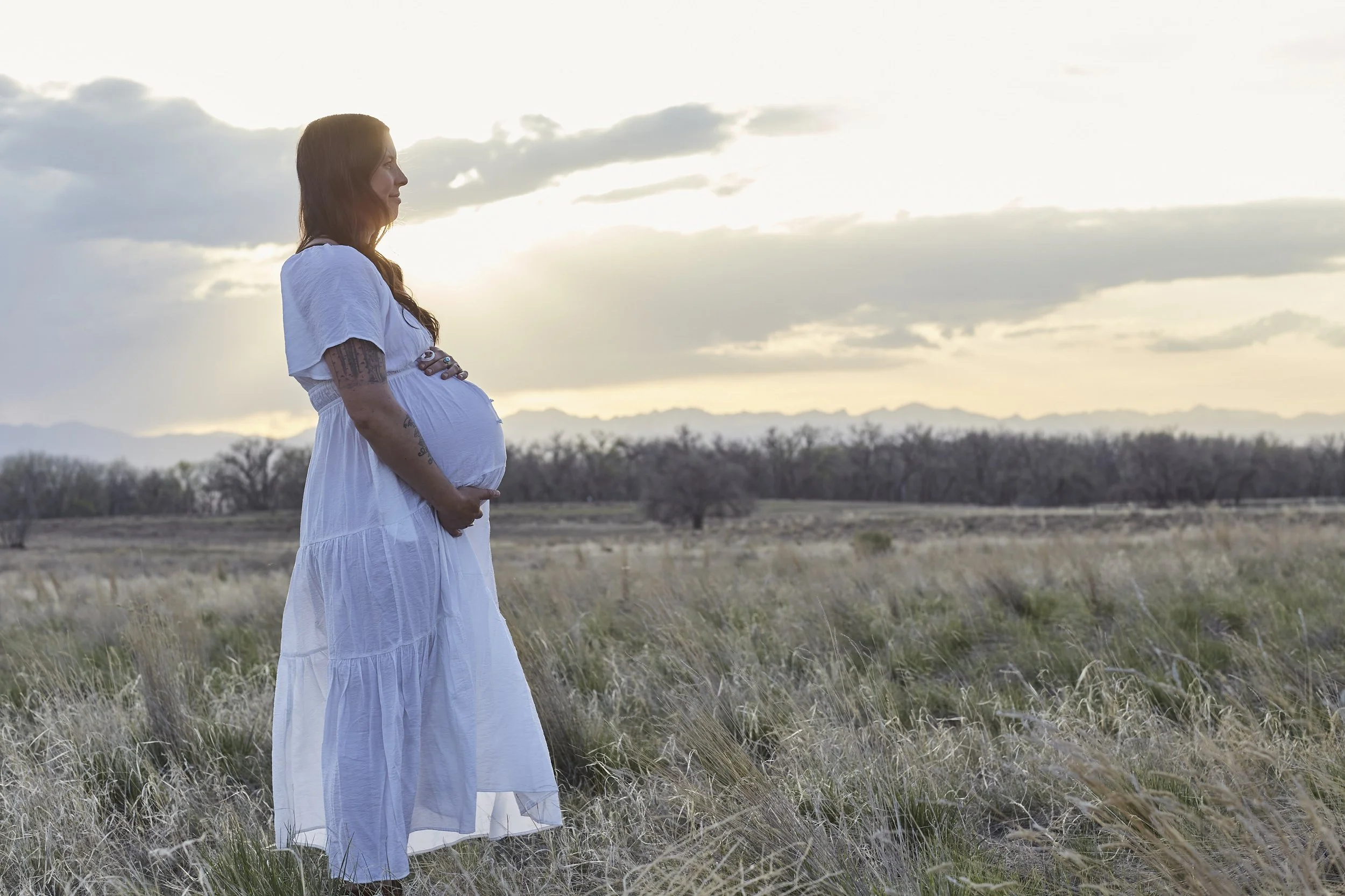 A pregnant woman wearing a white dress stands with her arms crossed over her belly in a grassy field during sunset.