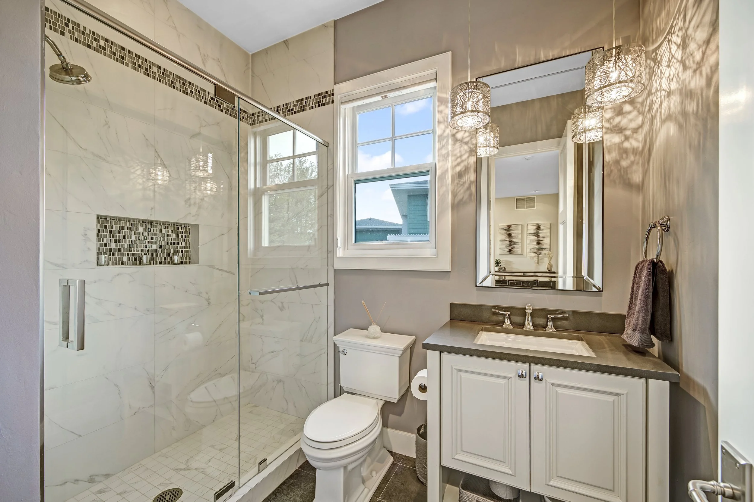 Bathroom with walk-in shower featuring white marble tiles and a decorative mosaic strip, three-pane window letting in natural light, mirror, white vanity with a sink, and a toilet, with modern pendant lights and a towel hanging nearby.