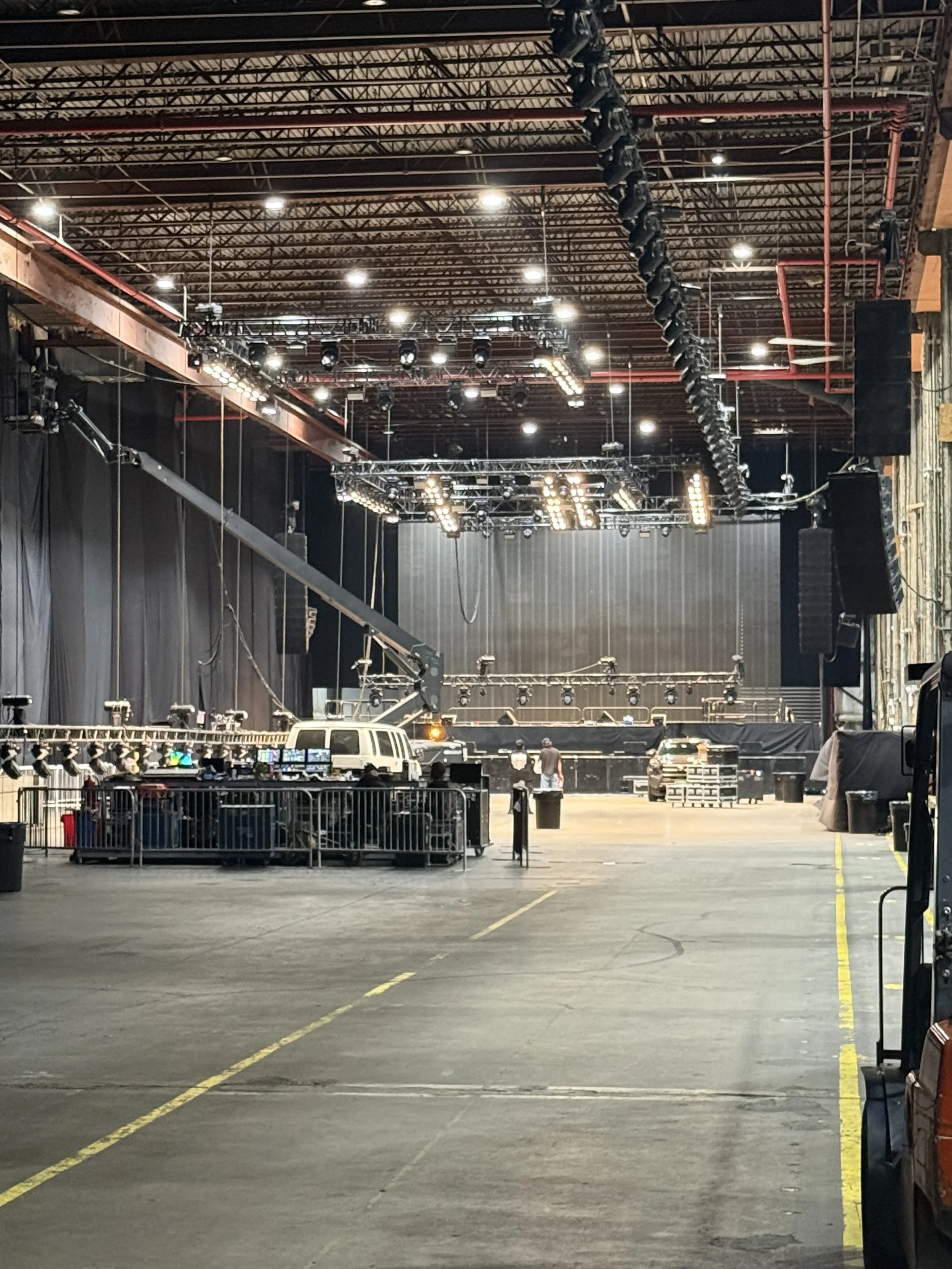 An indoor stage setup with lighting and sound equipment, a vehicle, and individuals working or setting up in a large spacious venue.
