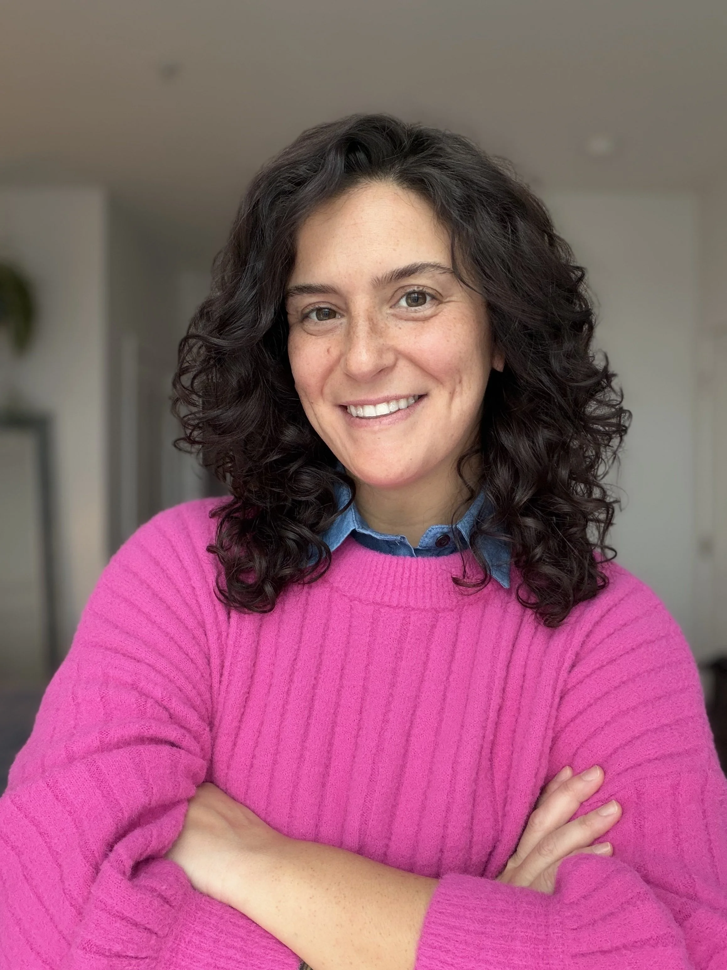 A woman with curly dark hair, wearing a blue collared shirt under a pink sweater, smiling with arms crossed.