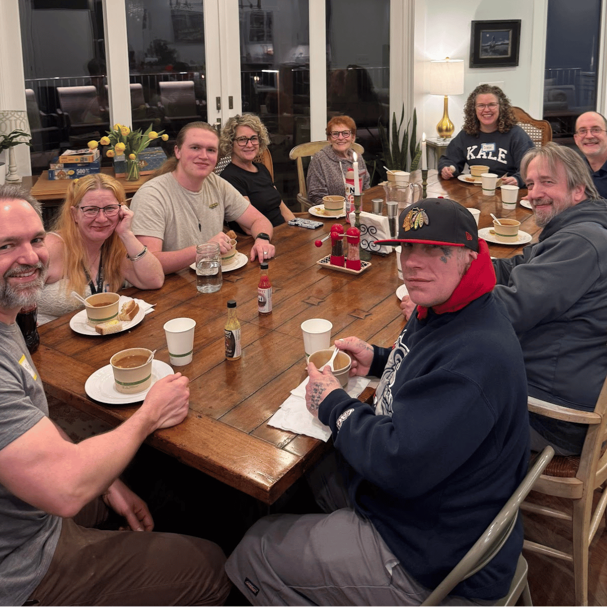 People gathered around a wooden dining table enjoying a meal, with some holding cups and others with plates of food, in a cozy indoor setting with large windows and lamps.