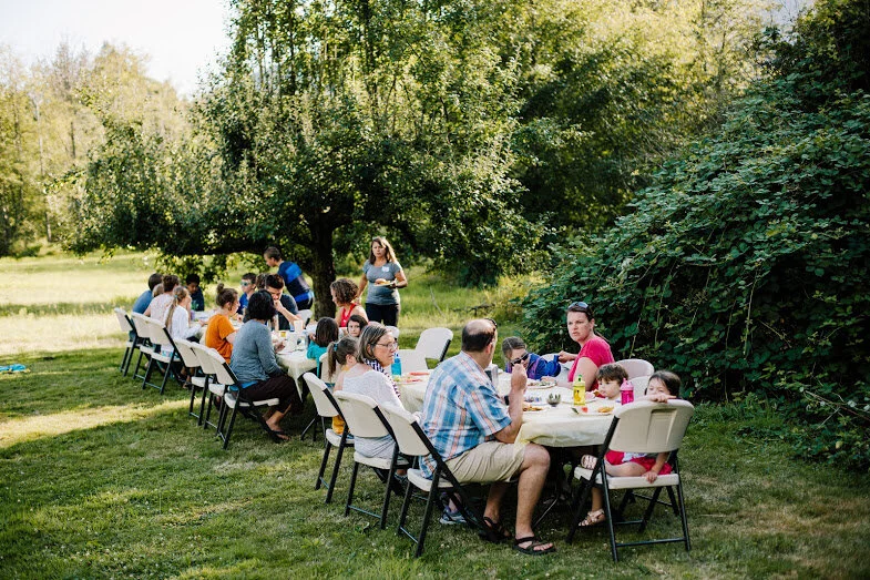 Family and friends gather for an outdoor picnic or potluck in a lush green park with trees, sitting at long tables with white chairs.