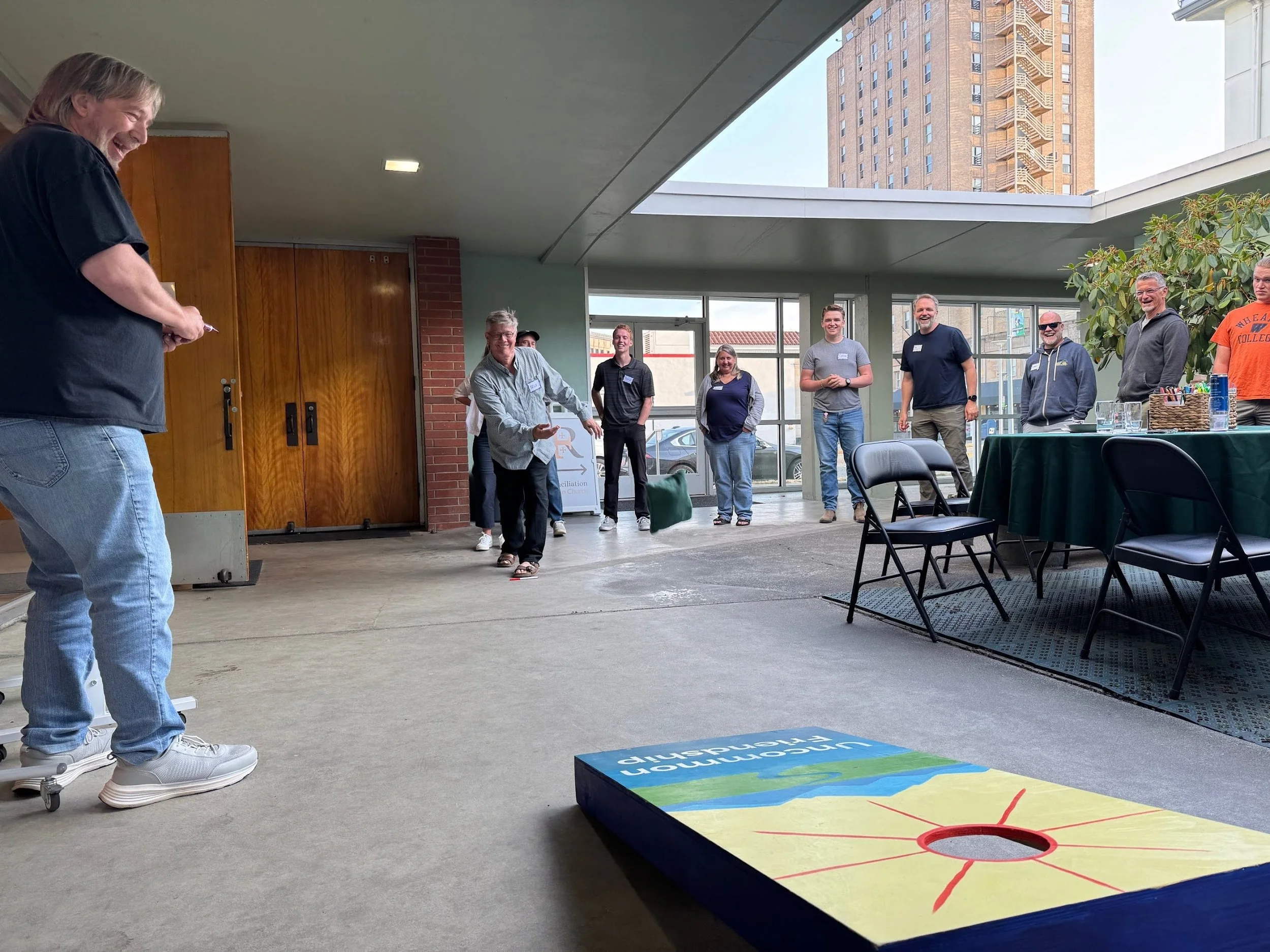 A group of people playing a bean bag toss game indoors, with some smiling and watching, and an urban cityscape visible through large windows.