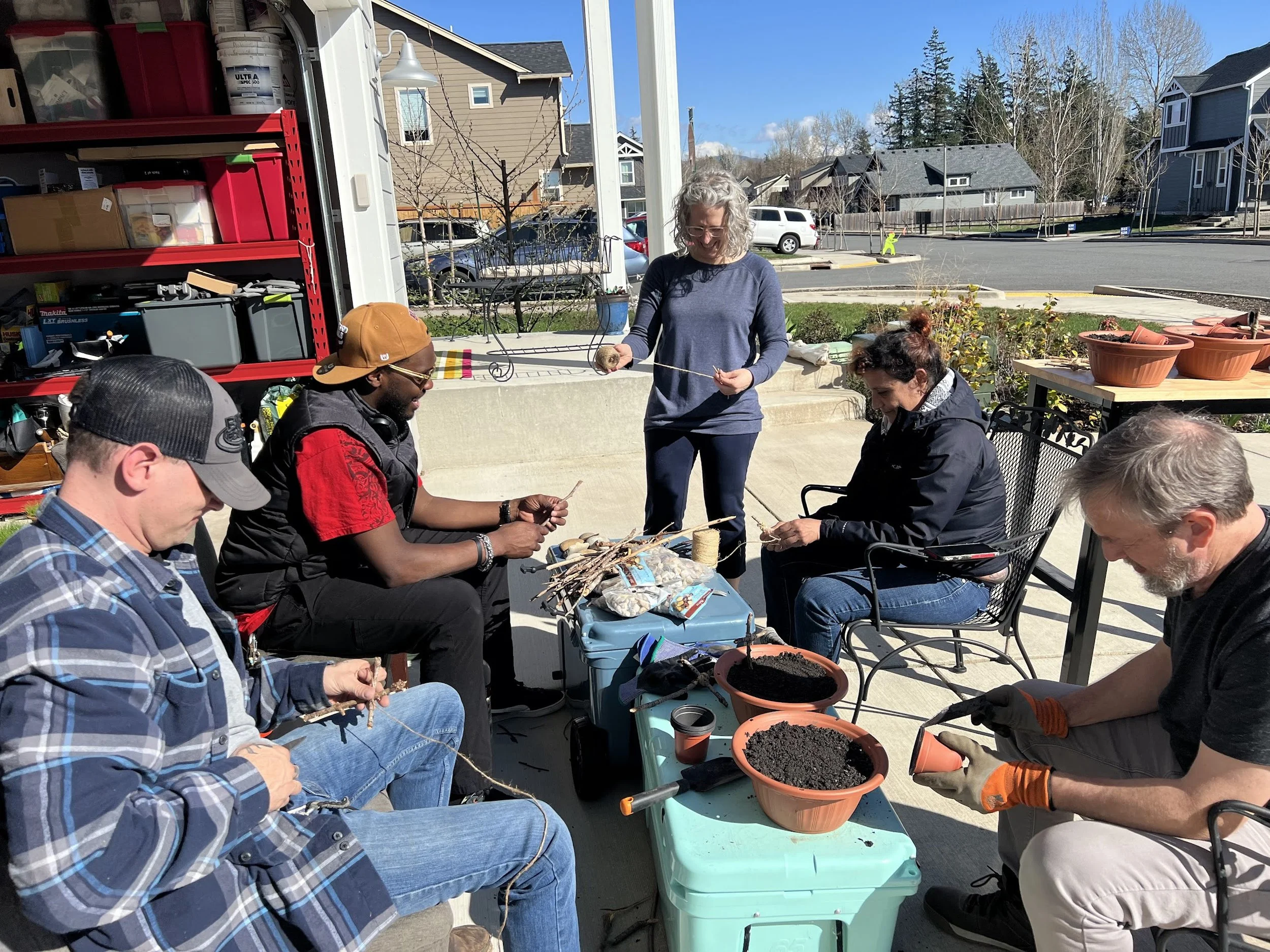 Group of people sitting outdoors, engaging in planting or gardening activities, with a woman standing and smiling, holding a small plant, surrounded by pots, soil, and gardening tools, with residential houses and trees in the background on a sunny day.