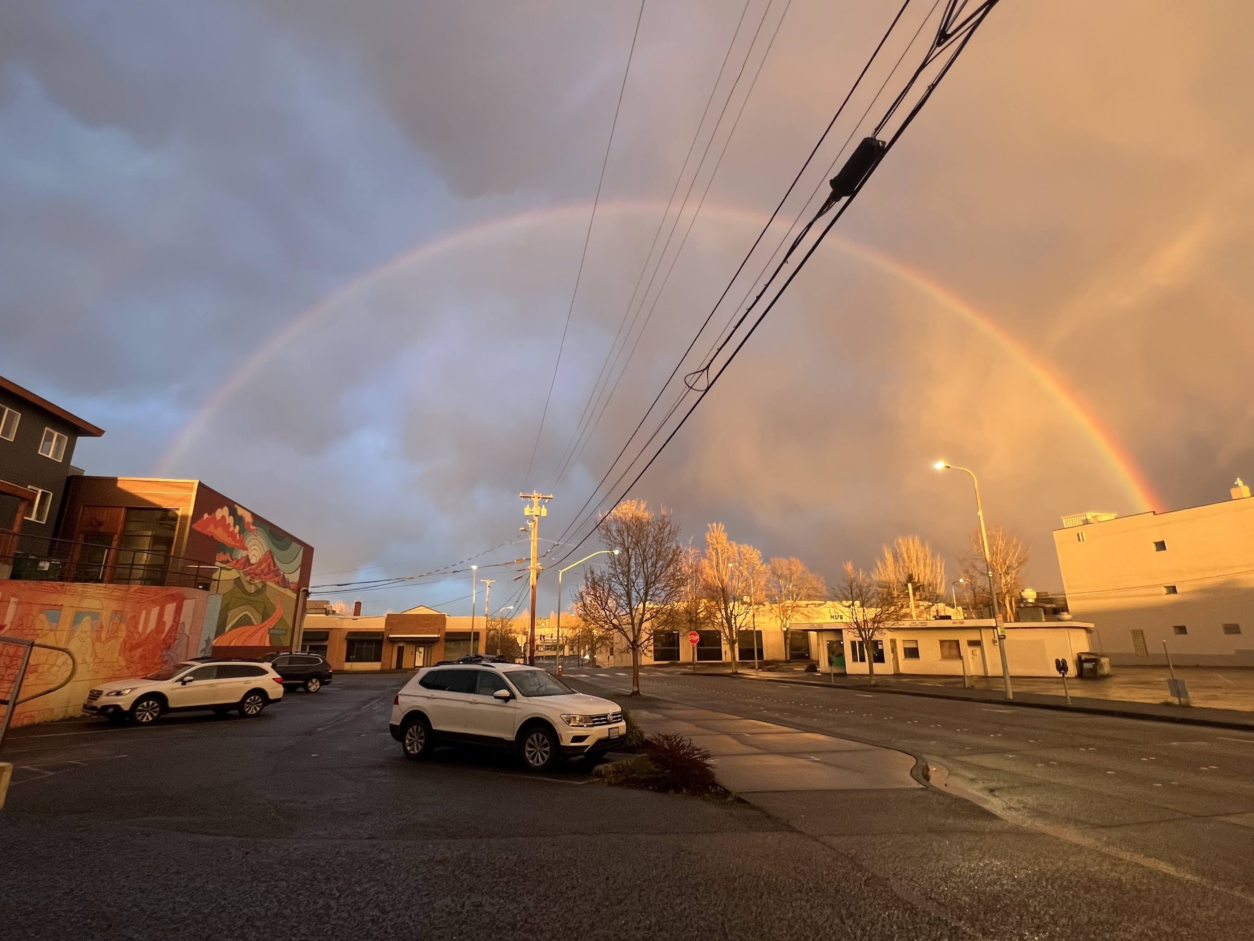 A parking lot during sunset with several cars, trees, and a building with a mural. A rainbow is visible in the sky, which has a mix of clouds and a warm glow from the setting sun.