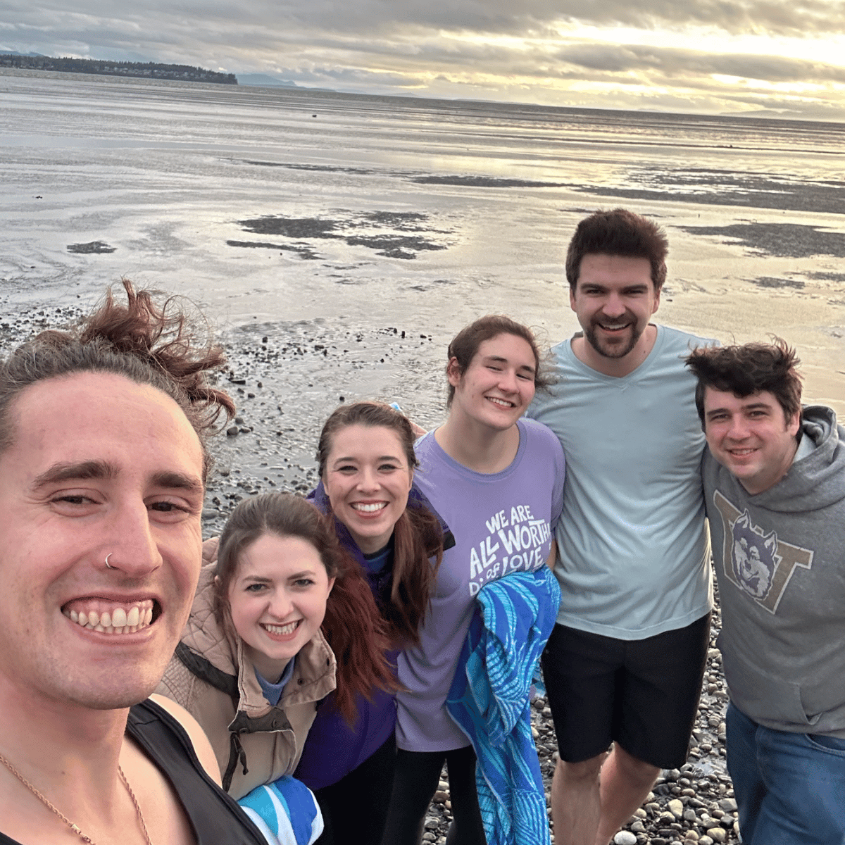 Group of six friends smiling on a rocky beach near the water during sunset, with overcast sky and distant shoreline in the background.