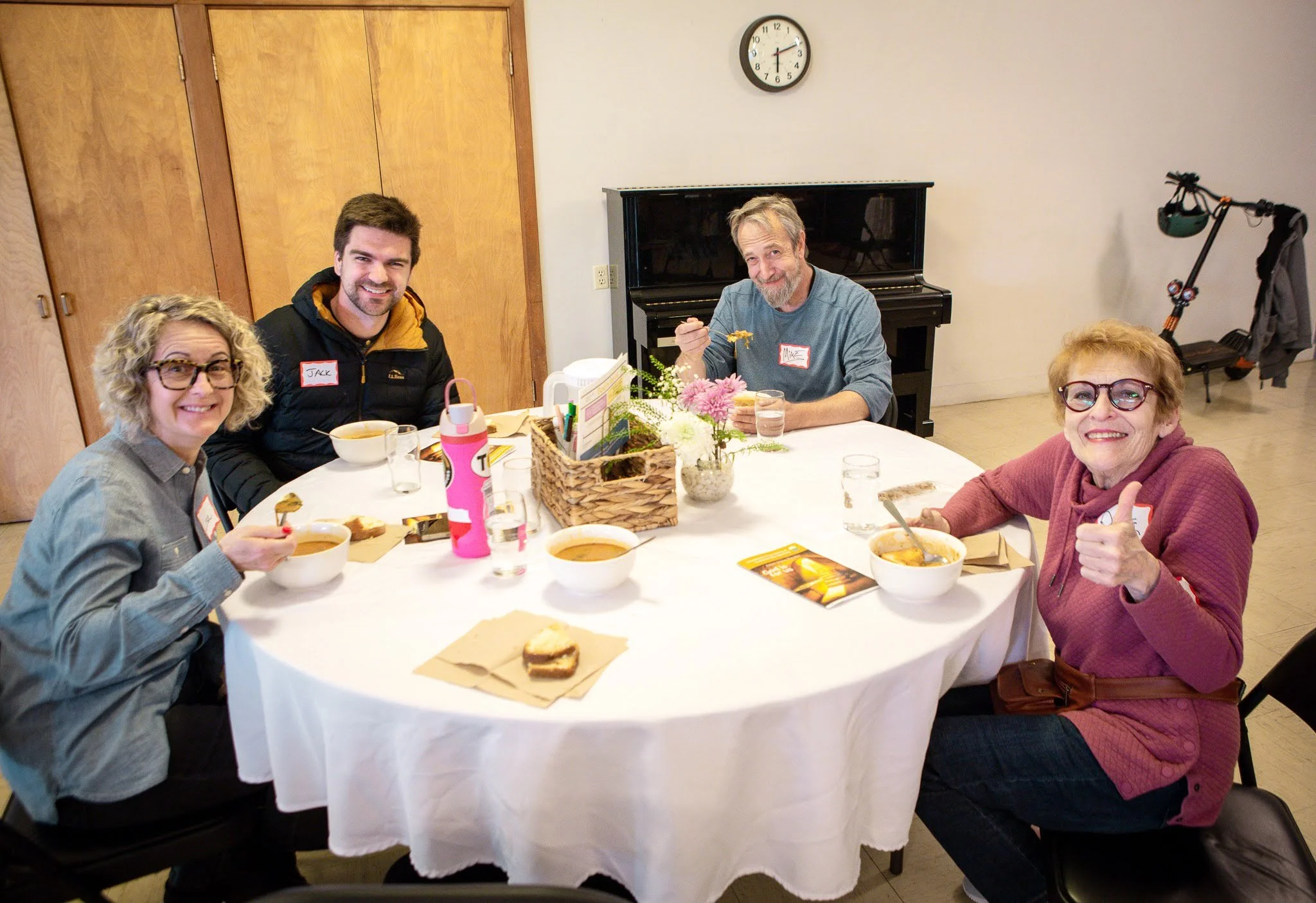 Four people sitting around a round table with white tablecloth having a meal. Three women and one man, all smiling. The woman on the left has curly blonde hair and glasses, the man has short dark hair and a beard, the man on the right has gray hair and a beard, and the woman on the far right has short red hair and glasses. There are bowls of soup, a basket of bread, and cups on the table. In the background, a black piano, a clock, and a scooter are visible.