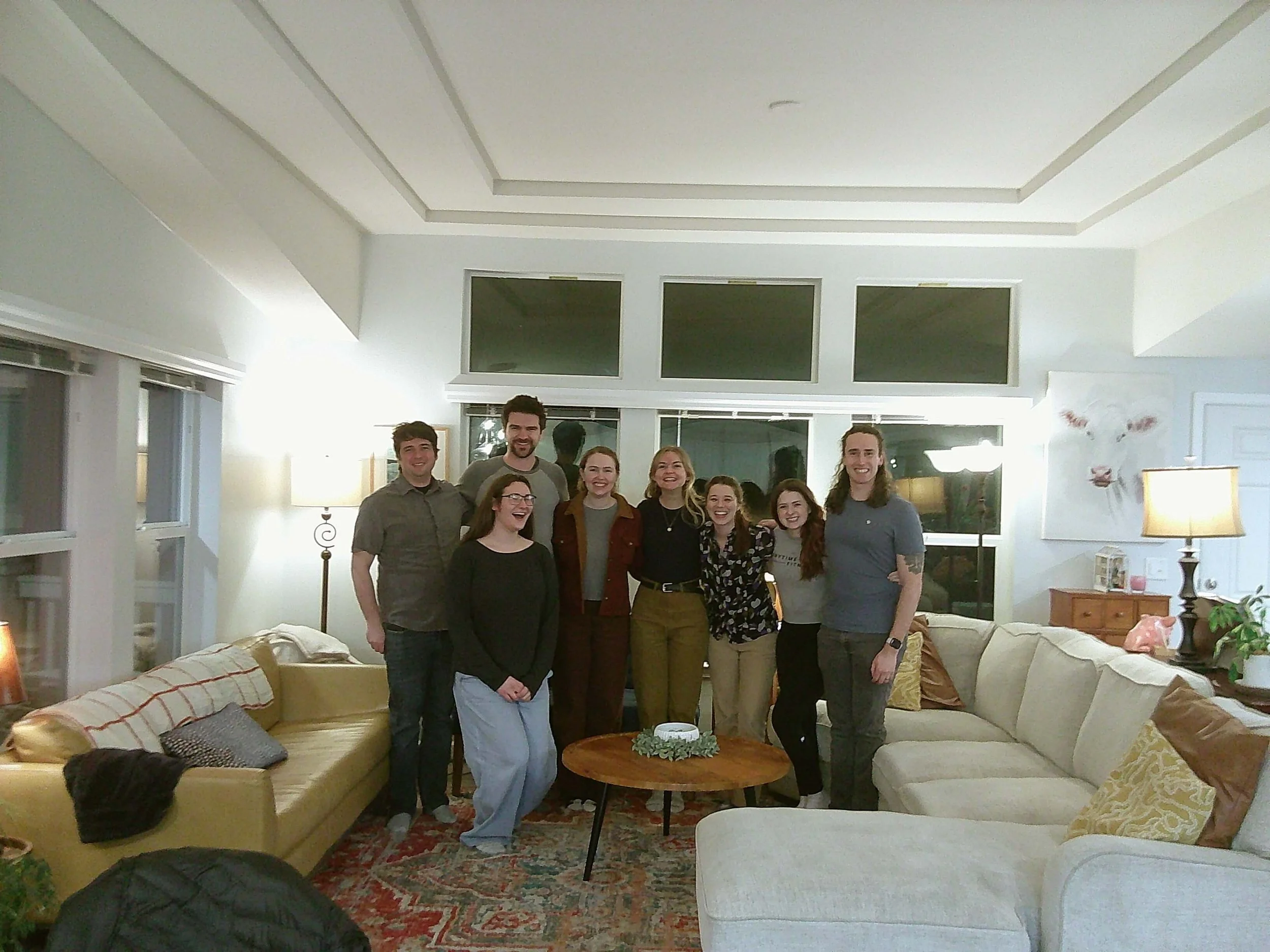 A group of nine people standing together in a living room, smiling at the camera. The room has a large cream-colored sectional sofa, a wooden coffee table with a decorative centerpiece, and windows with blinds. The background features a wall with three windows and a painting.