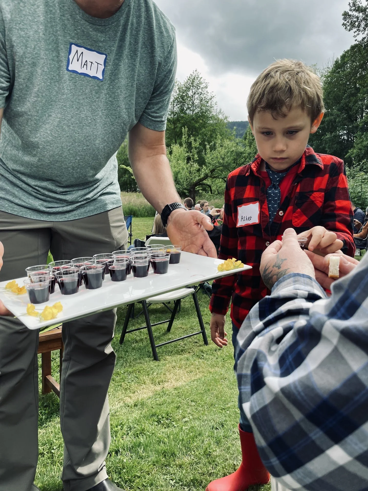 A young boy wearing a red and black plaid shirt and a name tag reading 'Asher' is standing outdoors during an event, reaching out towards a tray of small glasses filled with a dark red drink. An adult holding the tray has a name tag reading 'Matt.' Other people are visible in the background, sitting on chairs in a grassy area with trees and cloudy sky.