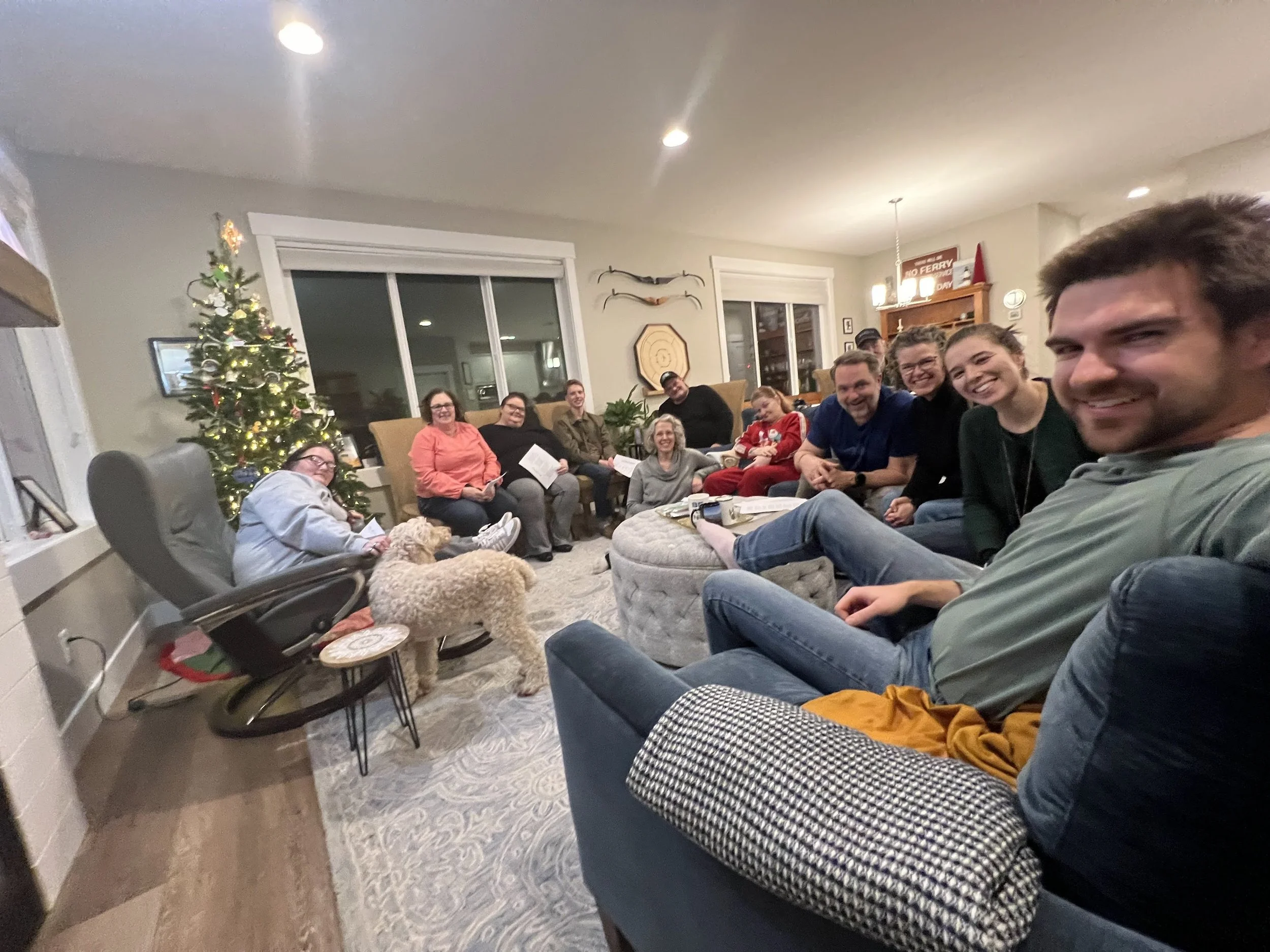A group of people sitting in a living room, smiling at the camera during a holiday gathering, decorated with a Christmas tree and festive decorations.
