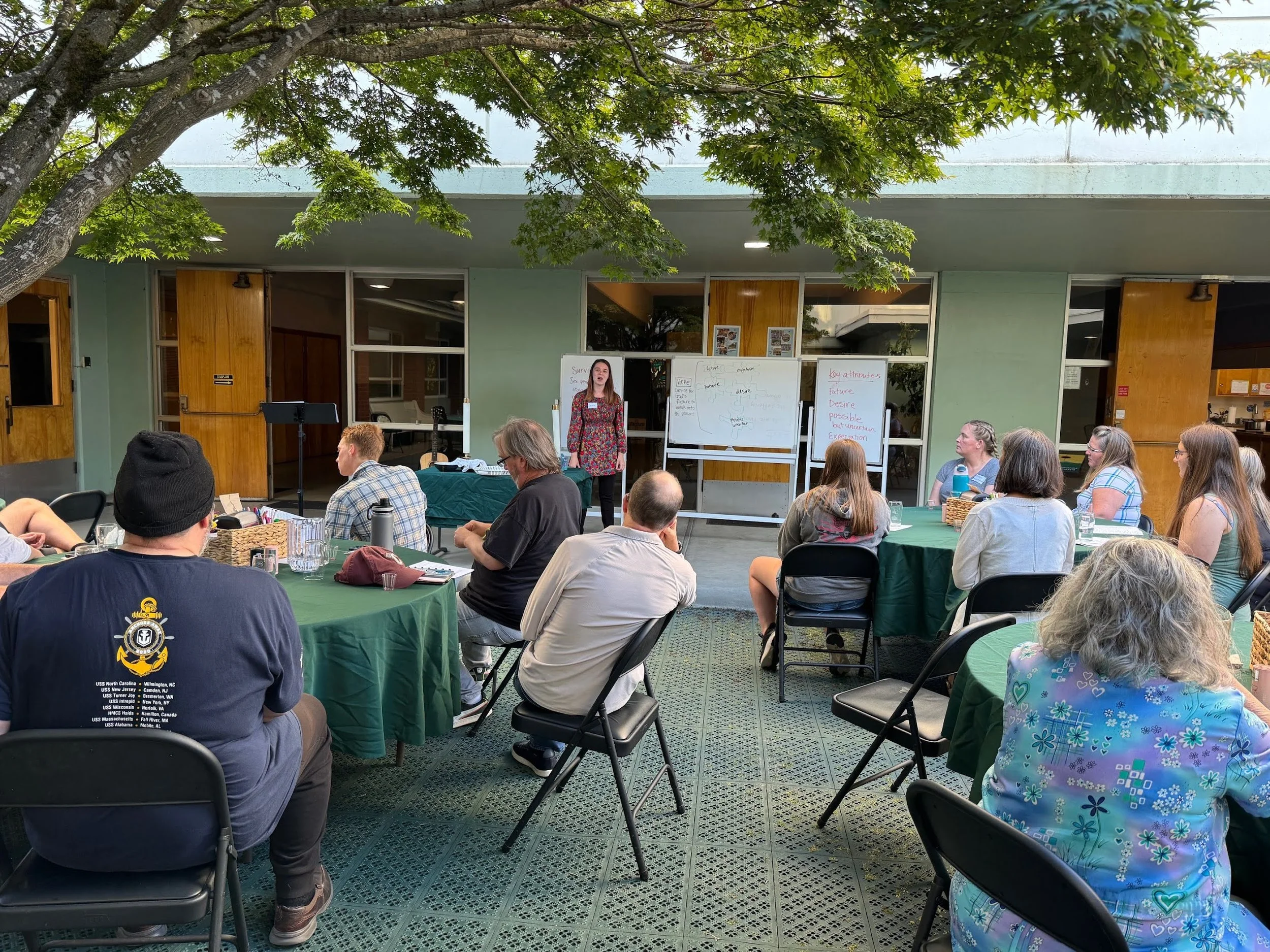 Outdoor classroom with people seated at green covered tables, a woman standing at the front near whiteboards, trees overhead, and a building in the background.