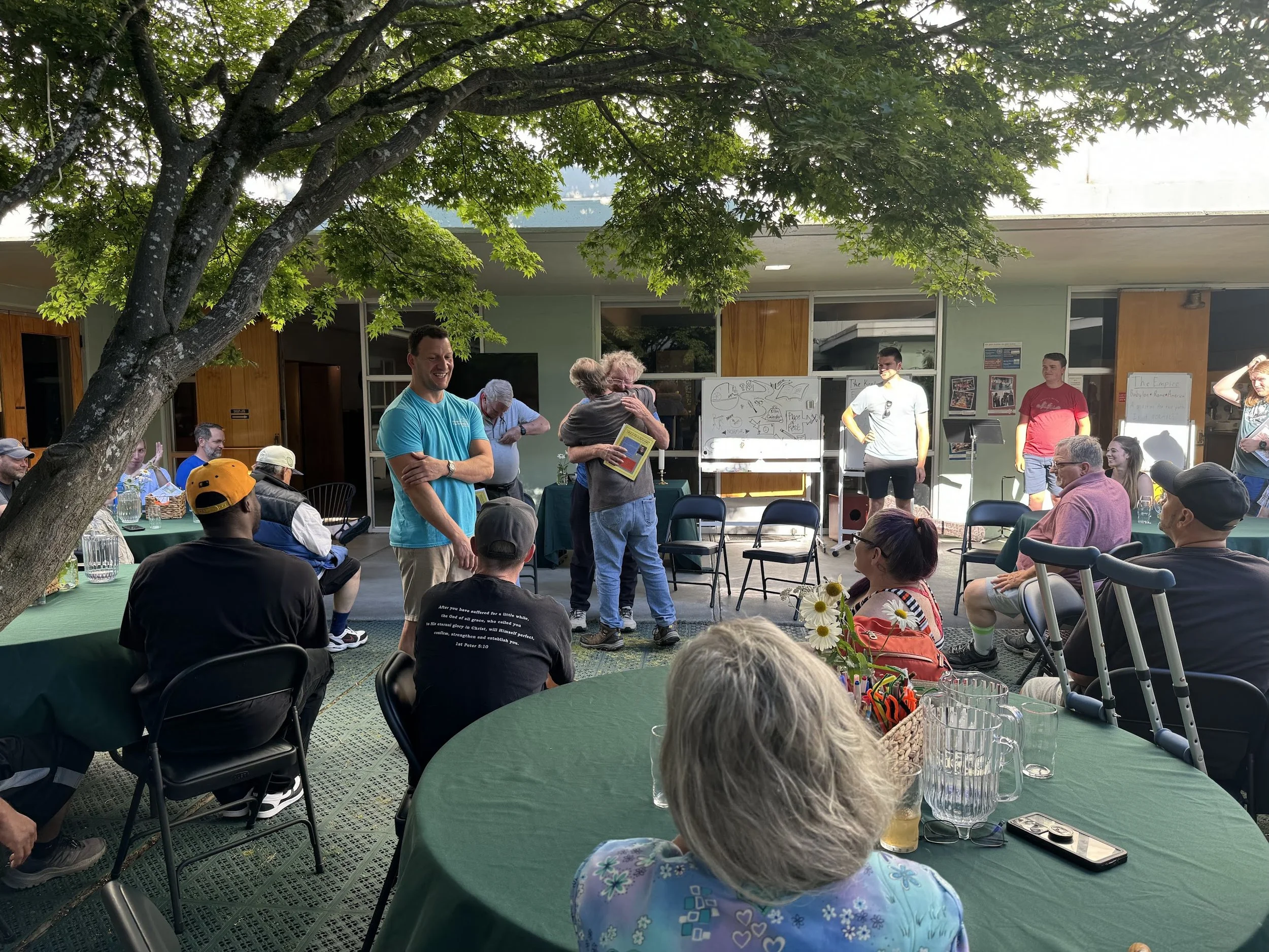 People attending an outdoor event under a tree, with some standing and others sitting at green-covered tables. There are whiteboards with drawings and text in the background, and the setting appears to be in a patio or courtyard area.