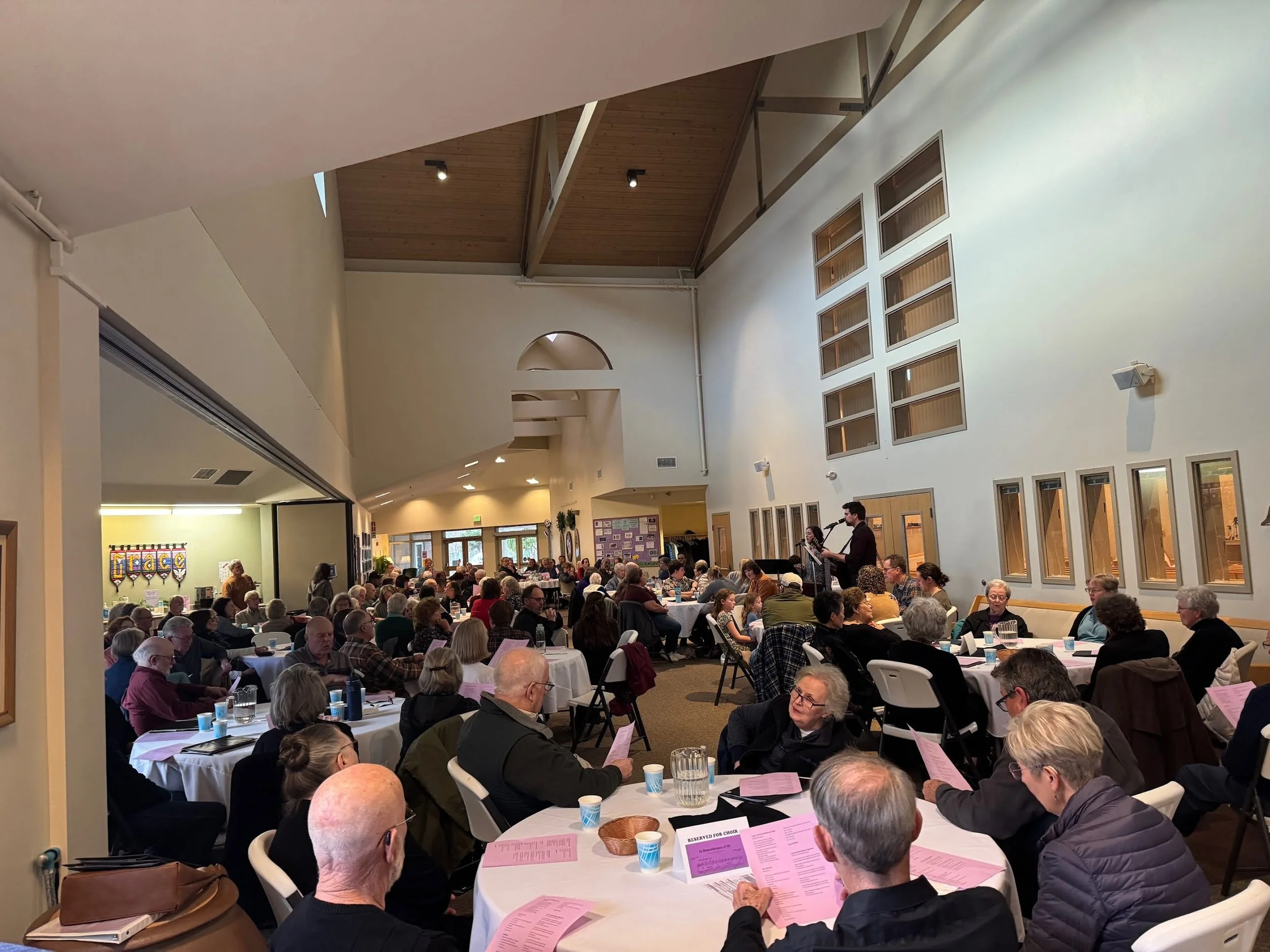 A large social gathering in a spacious room with high ceilings and wooden accents. Attendees sit at round tables covered with pink and white tablecloths, reading programs. A musician performs on stage, and people appear engaged.