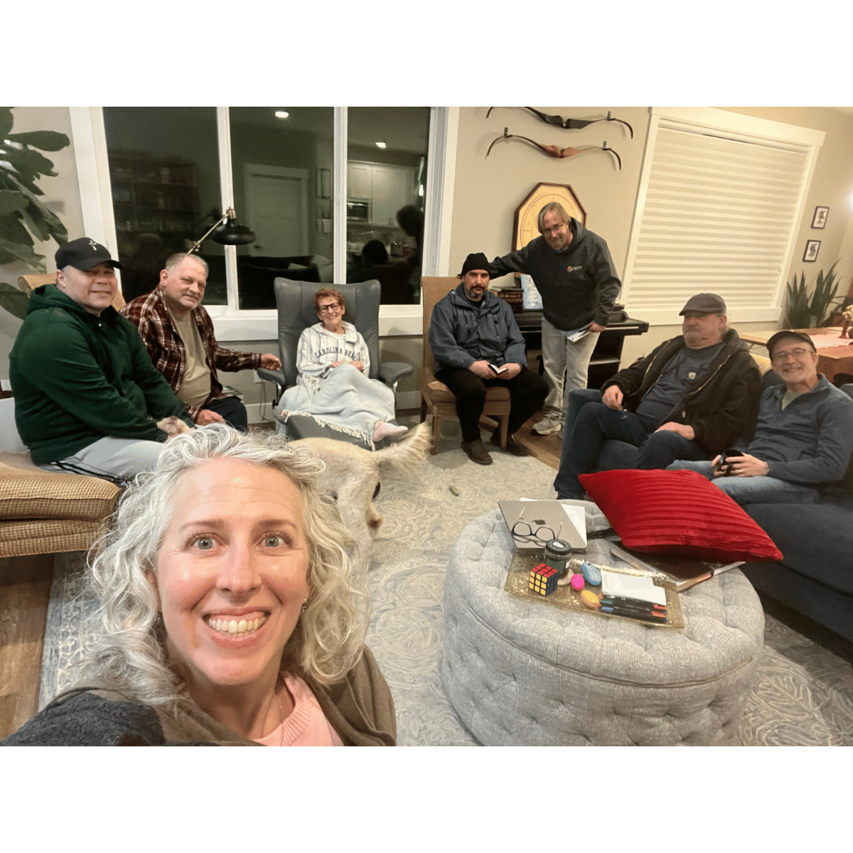 A group of nine people gathered in a living room, sitting on various chairs and couches, with one person taking a selfie in the foreground. There is a woman with curly blonde hair smiling at the camera, and a large dog sitting on the floor. The group appears to be enjoying each other's company in a casual setting, with various personal belongings and decorative items visible around them.