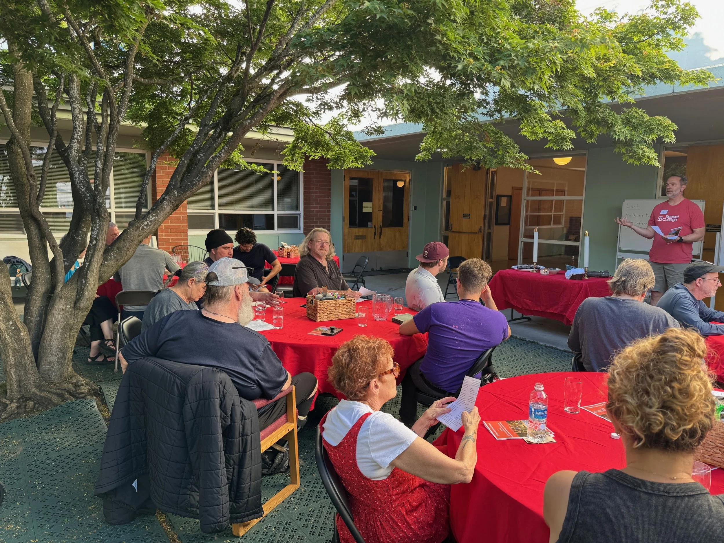 People gathered outdoors around tables with red tablecloths listening to a man giving a presentation in front of a building with windows and a blue roof, under a large tree.