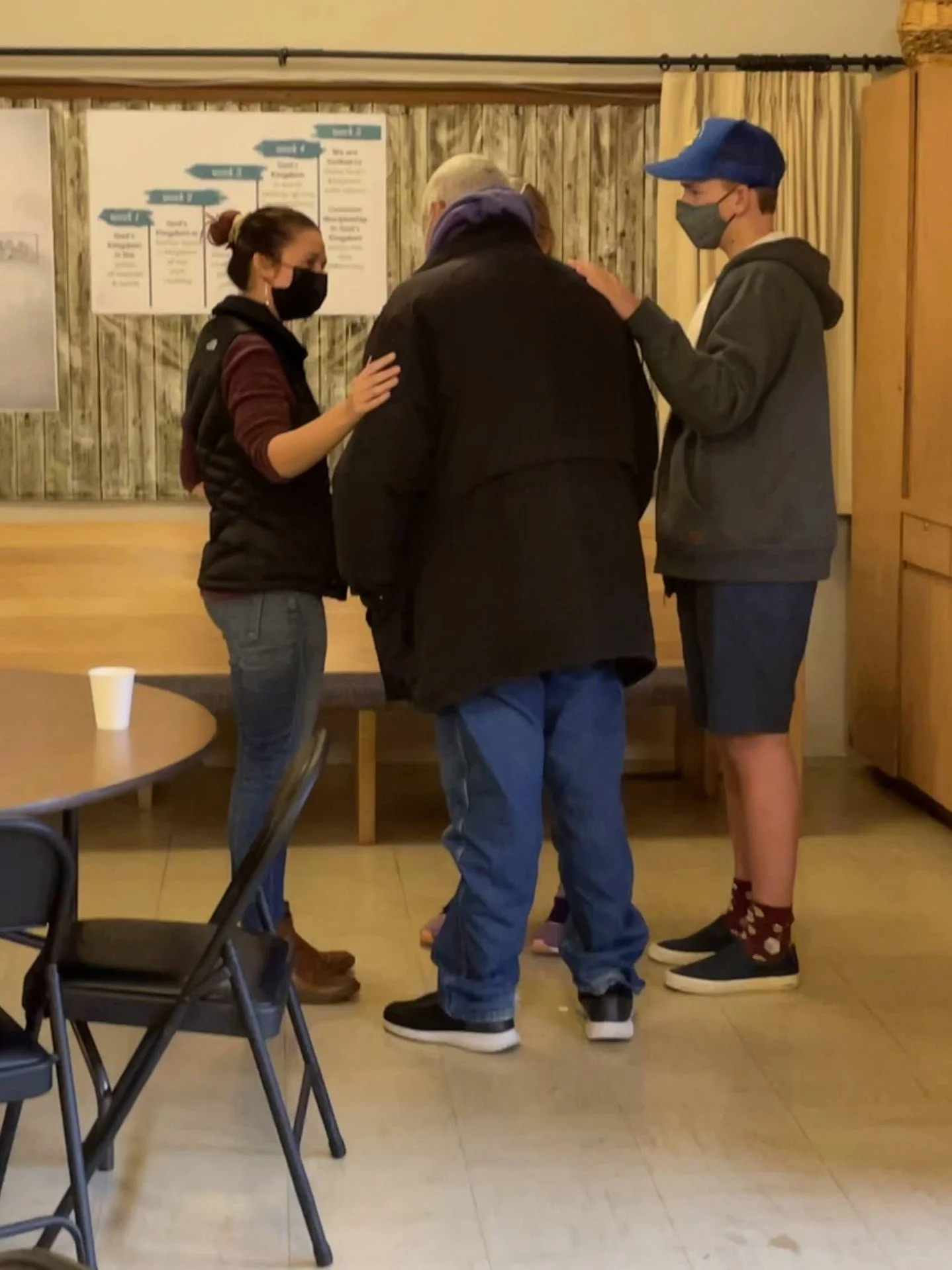 Three people are standing in a room, engaged in a conversation. One is a woman with a black face mask, the man has a bald head with a purple scarf, and the young man is wearing a baseball cap and a face mask. The room has wooden paneling on the walls, with a chart or informational poster in the background, and empty chairs and tables are visible in the foreground.