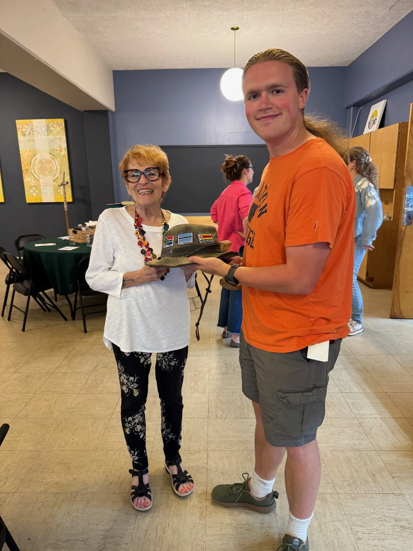 An elderly woman with glasses and colorful necklace smiles as she presents a hat with flags and patches to a young man in an orange T-shirt, in a room with blue walls and other people in the background.