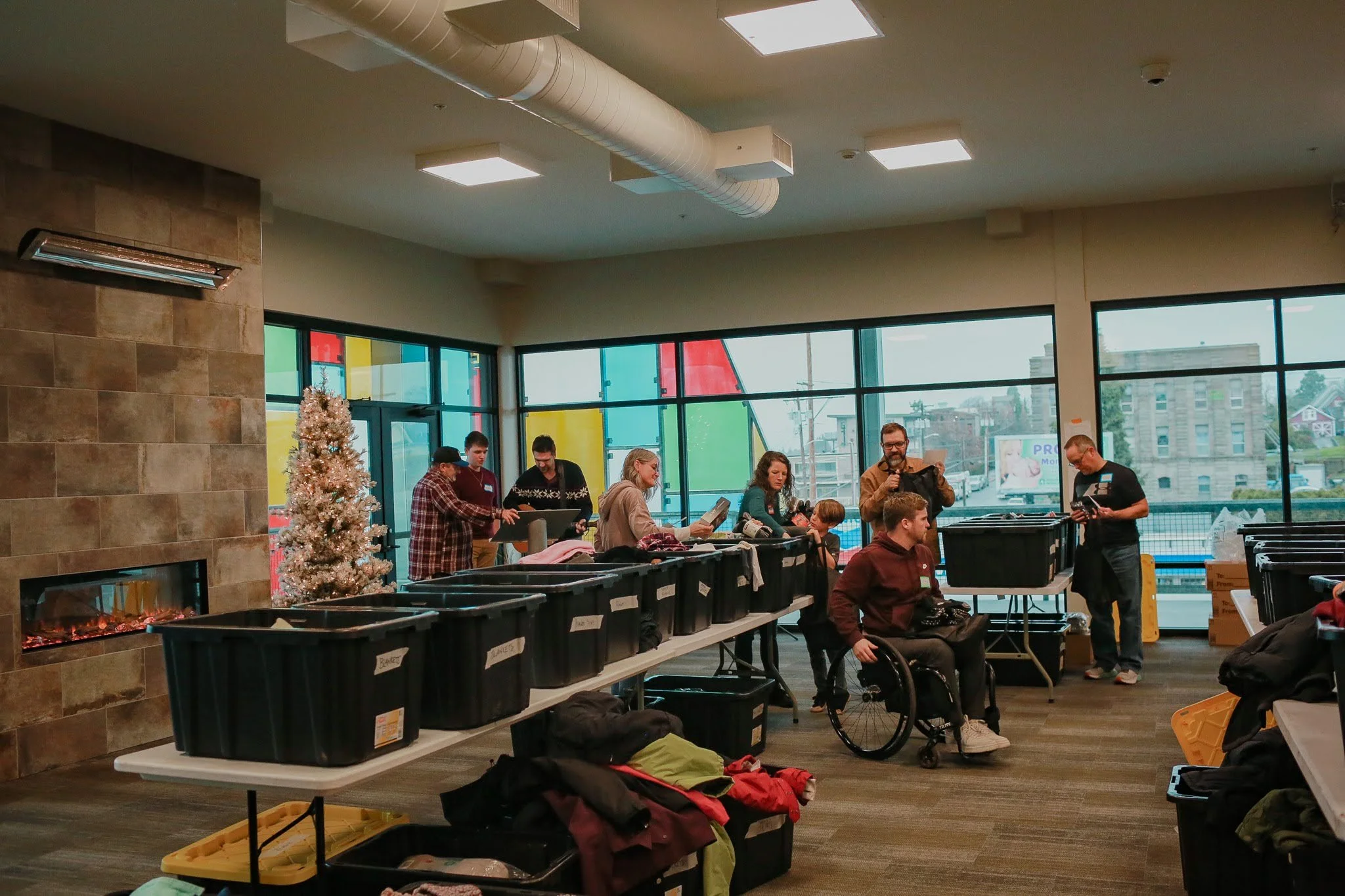 Indoor scene showing people gathered around tables with bins of clothing, a Christmas tree, and a fireplace, with large windows and a colorful mural outside.