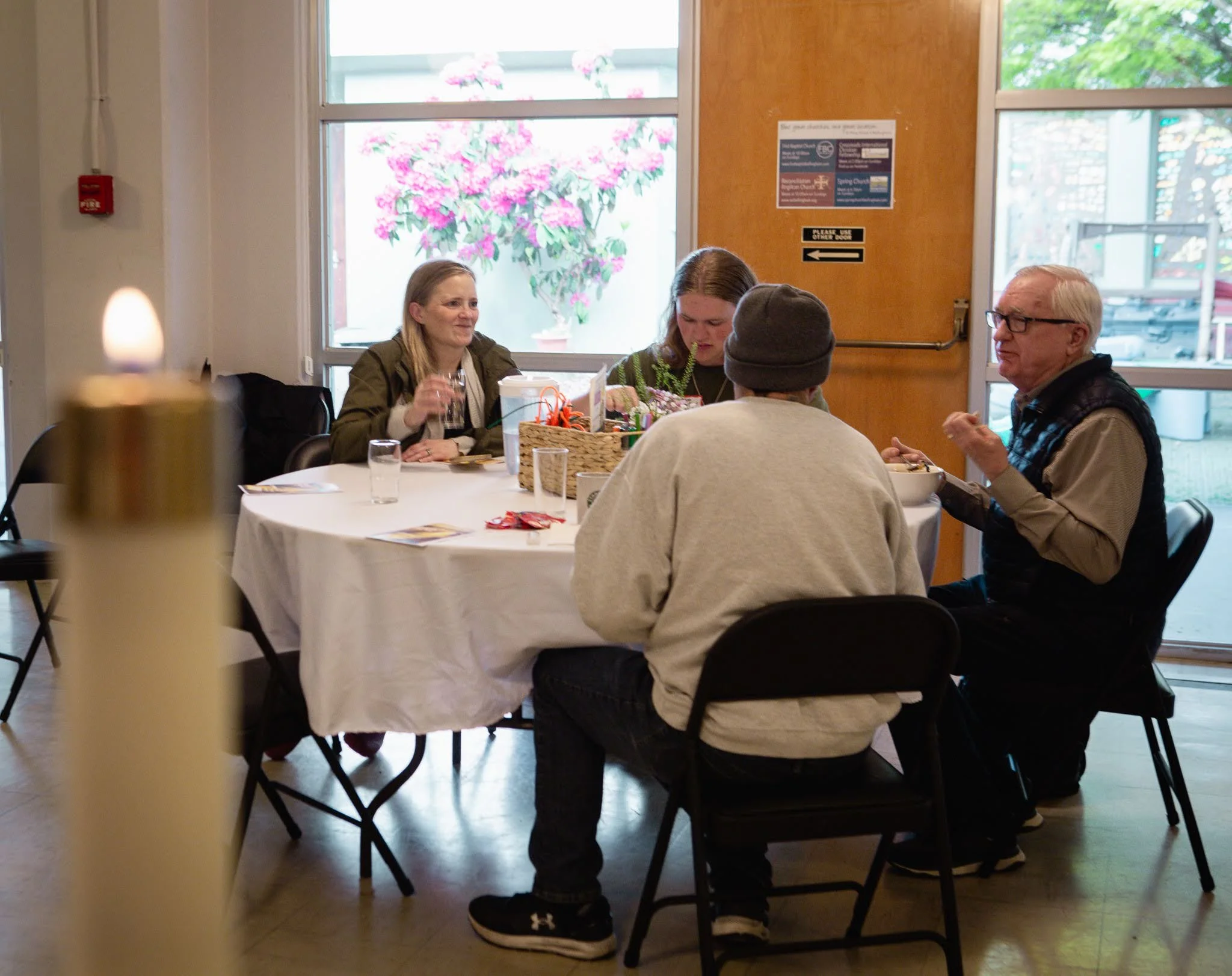 Four people sitting around a table in a room with large windows, with pink flowers outside, engaging in conversation and eating food.