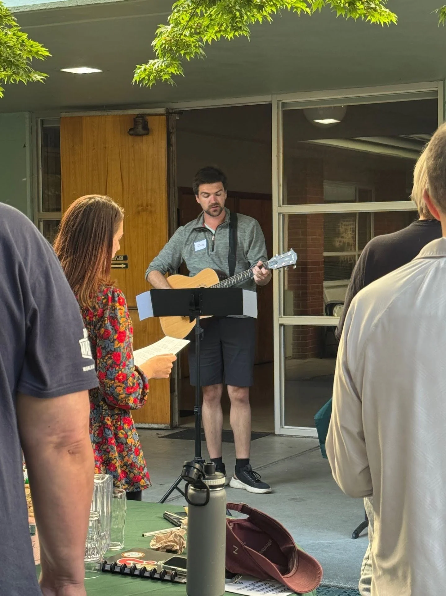A man is playing guitar and singing outdoors in front of a small audience. The audience members are standing, and some are holding papers. The setting appears to be outside a building with large windows and green tree foliage overhead.