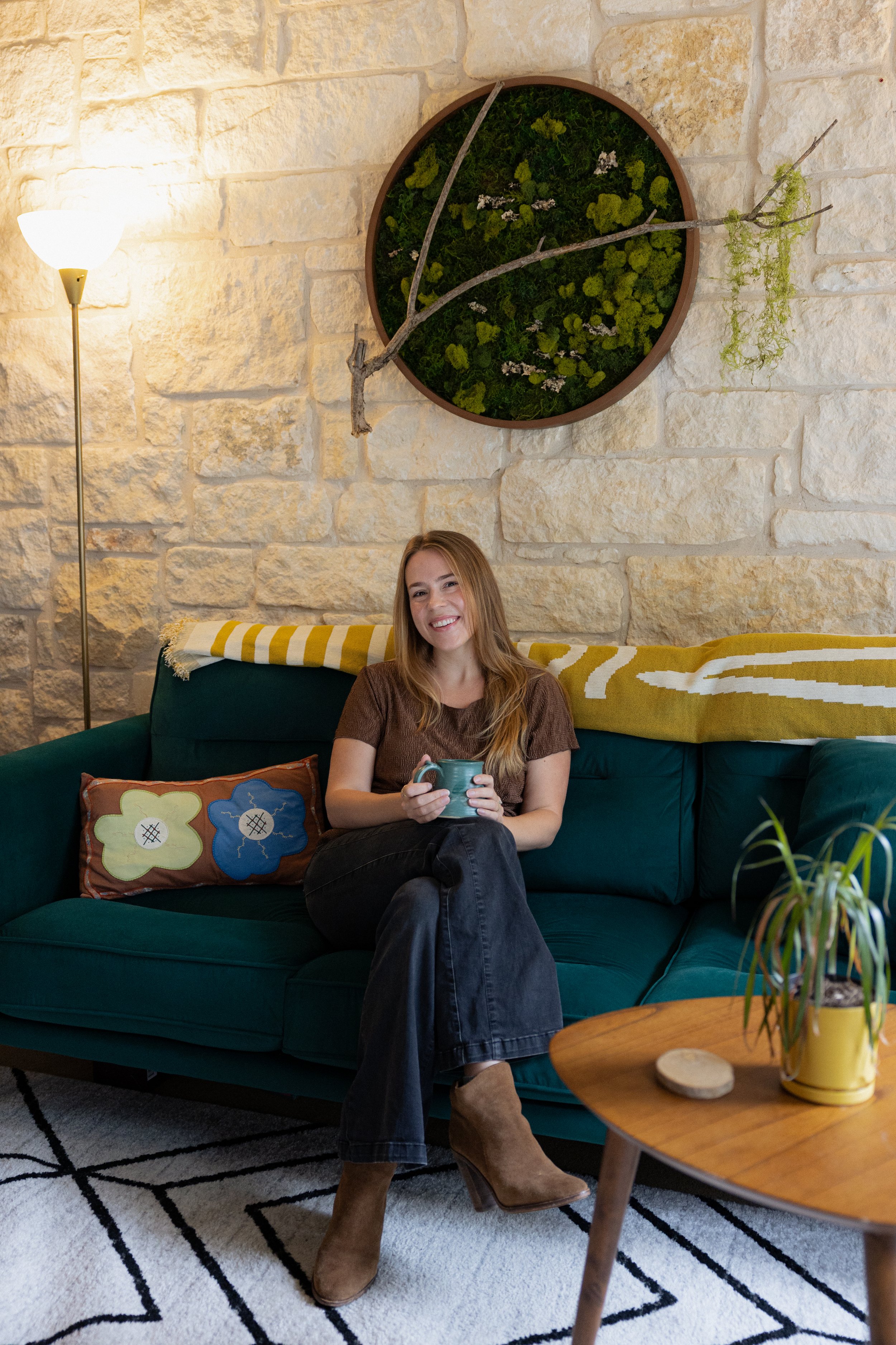 Marilyn is seated on a green couch with a yellow blanket across the back & a brown throw pillow with a green and blue flower. She is smiling at the camera, with both hands holding a coffee cup, resting on top of her knee.