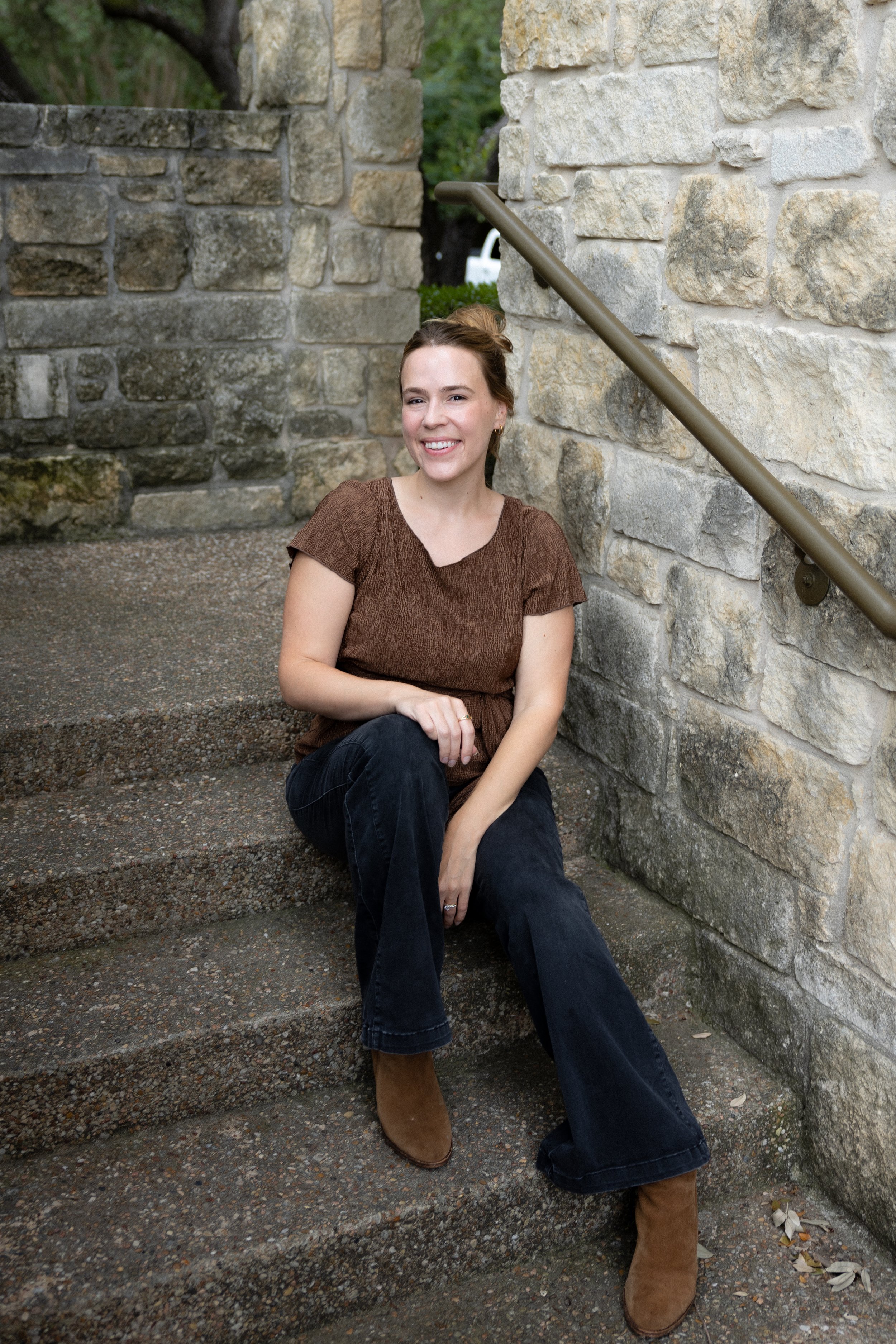 Marilyn is smiling at the camera, seated on stone steps with stone walls around her and a handrail on the right. She wears a brown shirt and shoes, dark pants, and her hair is pulled back.