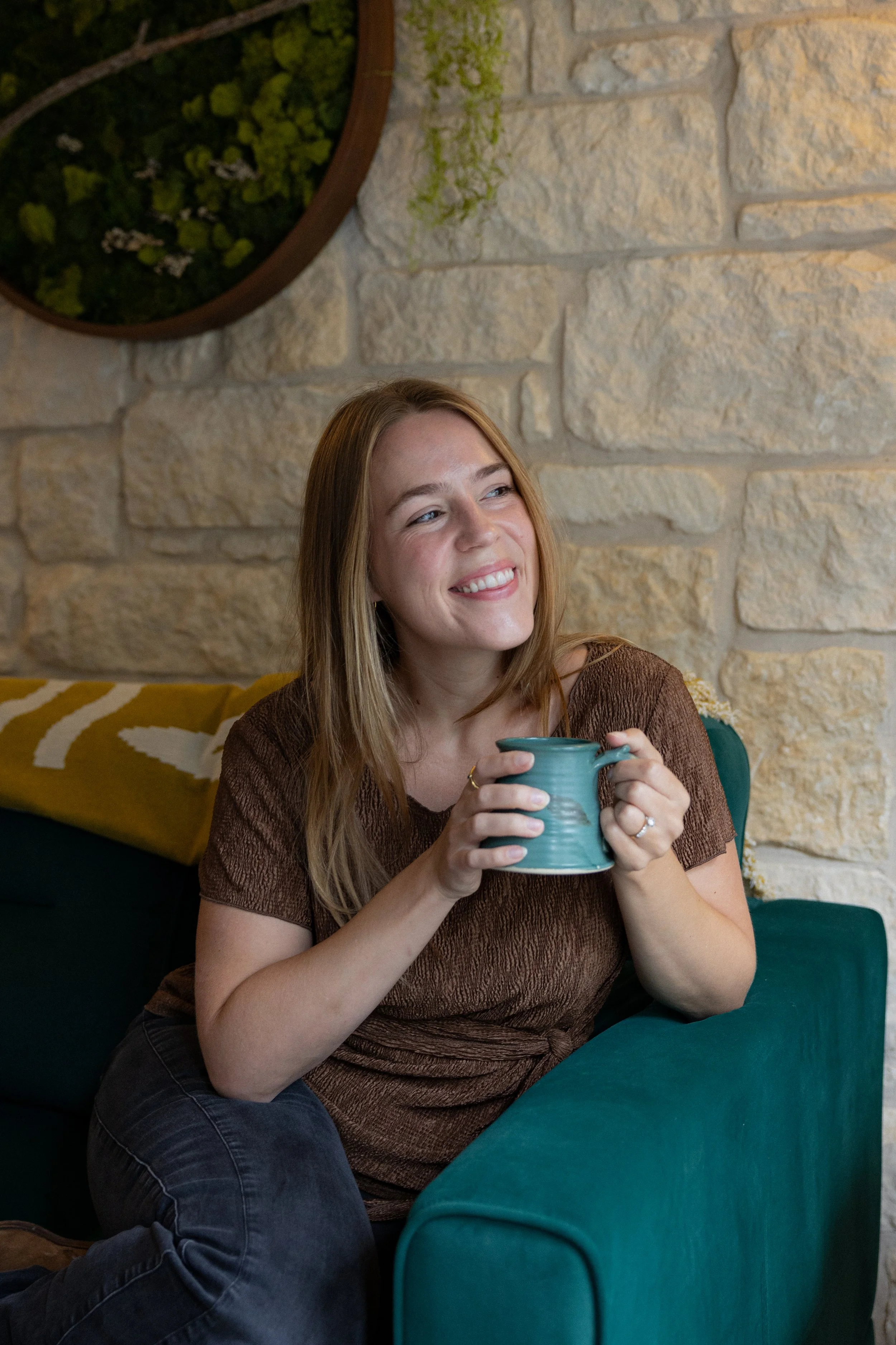 Marilyn smiles as she looks off to the right. She is wearing a brown shirt and holding a blue coffee mug with both hands. Her long, dark blonde hair is down, she is seated on a dark green couch, and a white brick/stone wall is behind her.