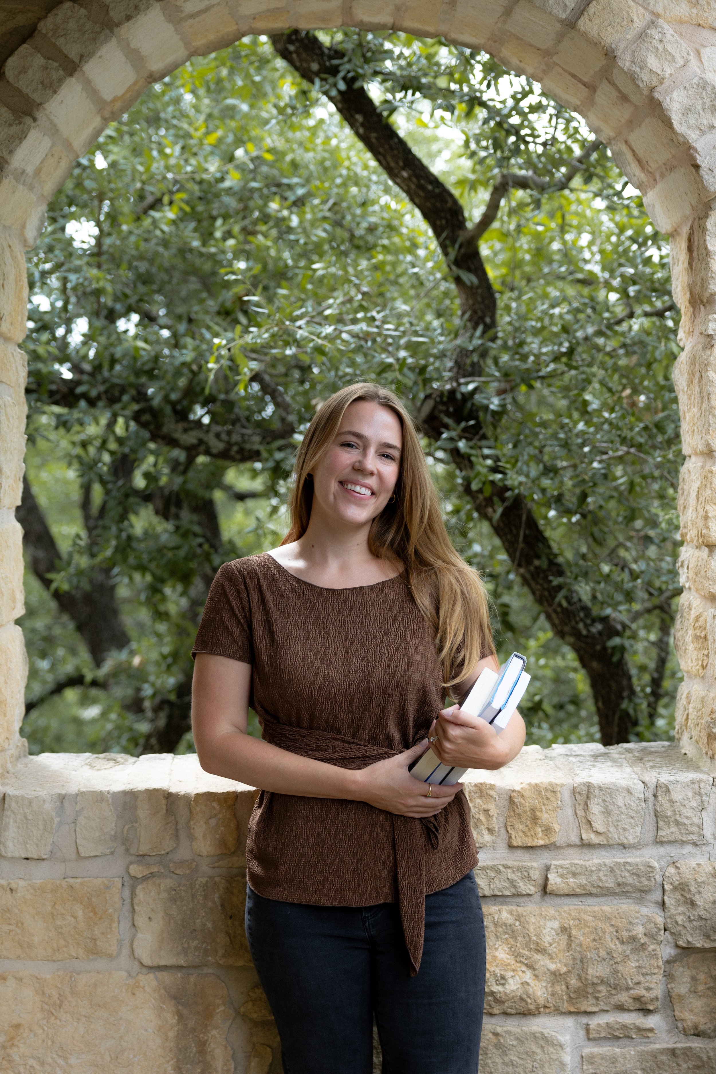 Marilyn smiles at the camera, standing in front of a stone wall with green trees in the background. She holds books, is wearing a brwown shirt, and her dark blonde hair is down.