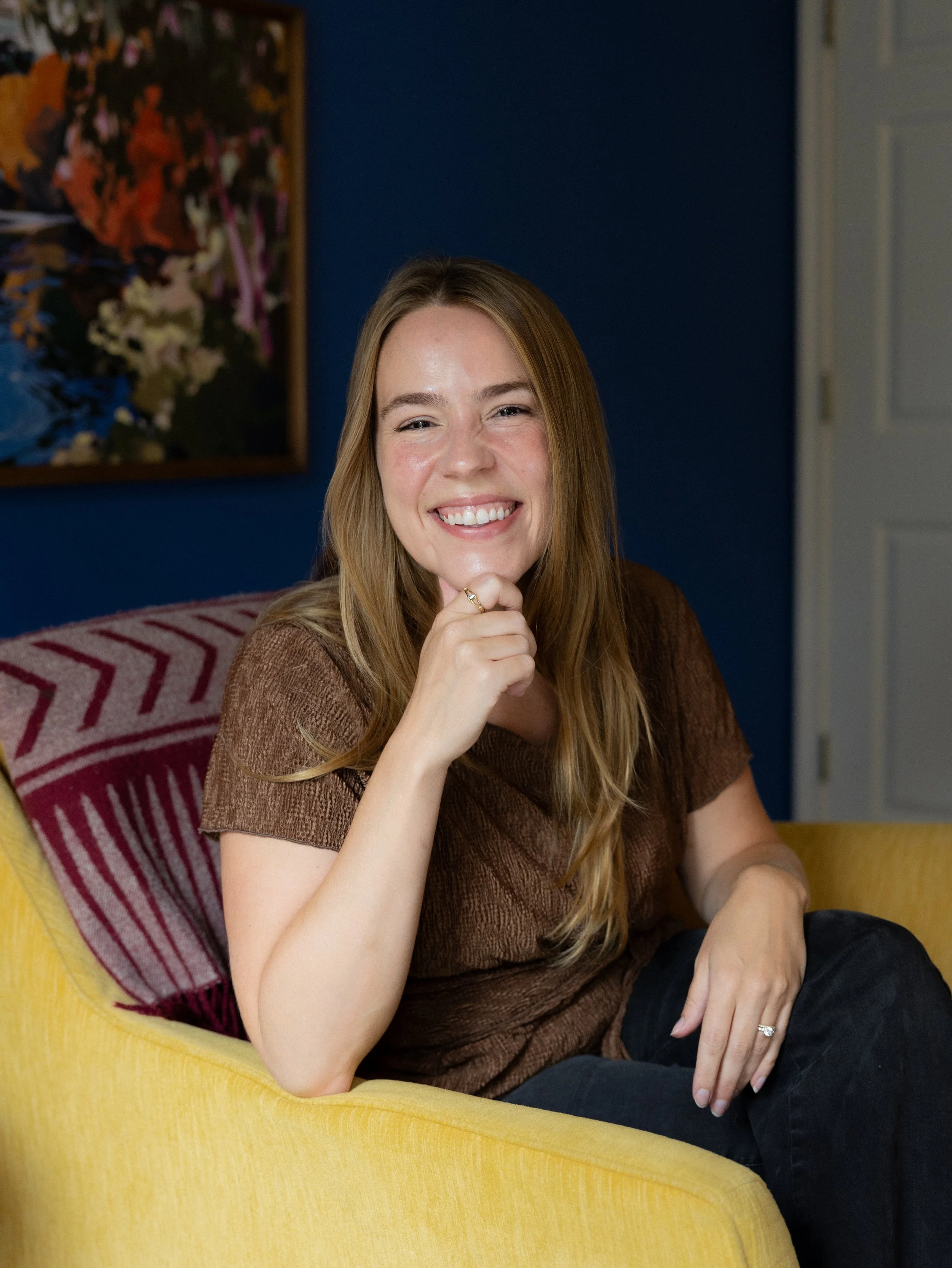 Marilyn smiles at the camera with her chin resting on her hand, and her hand propped up on her sofa. Her shirt is brown and her hair is long, dark blonde, and the background is dark blue with miscellaneous decor.