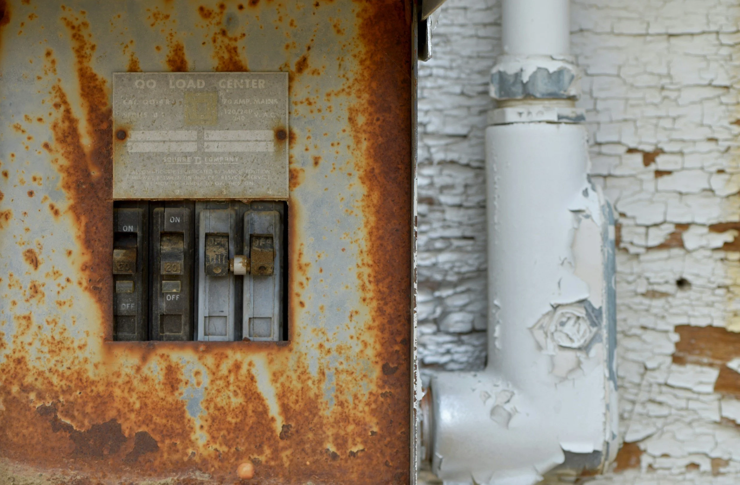 A rusty electrical load center with peeling paint and a background of weathered white brick wall and chipped paint pipe.