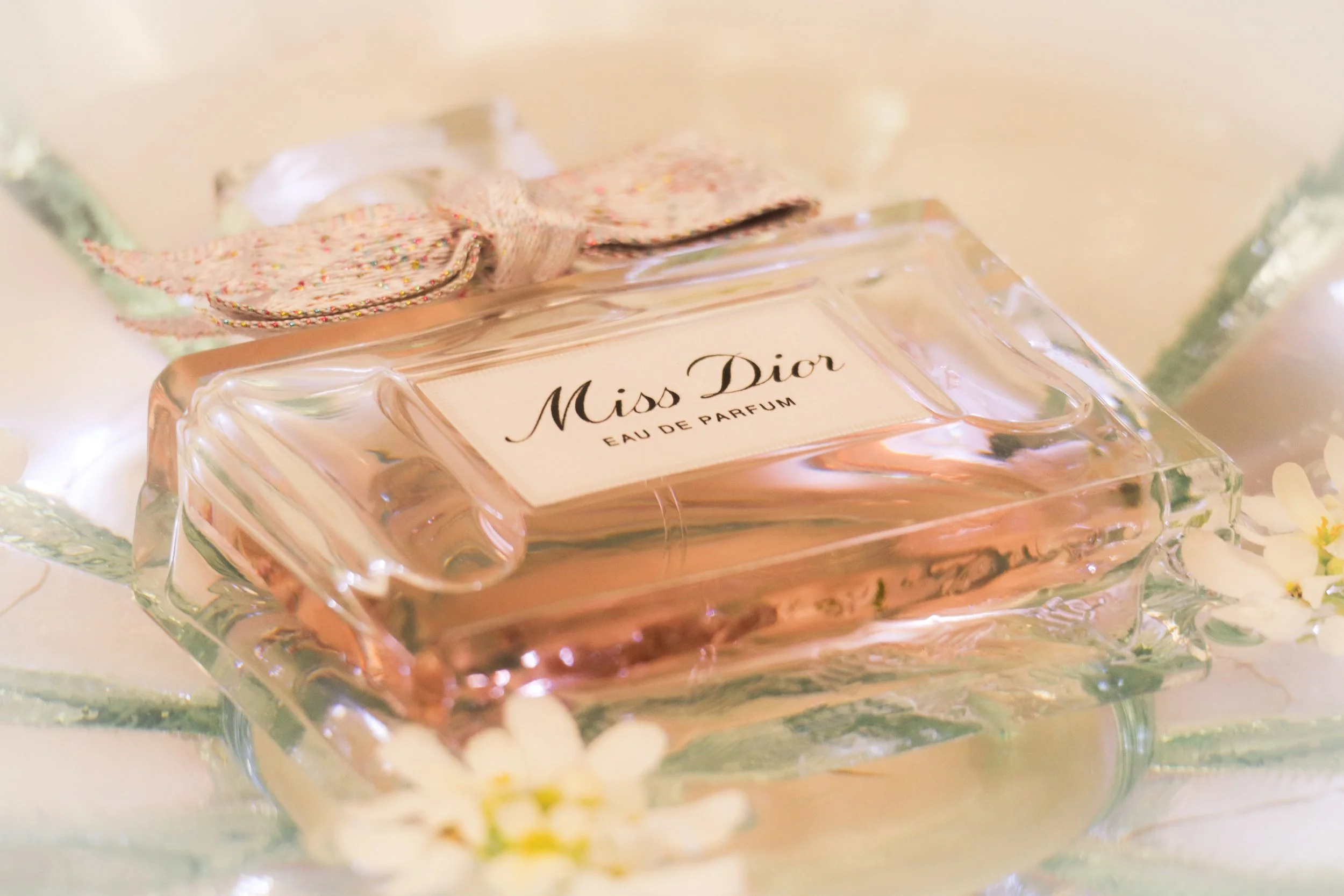 Flacon de parfum Miss Dior rose transparent avec un ruban en tissu fleuri sur le dessus, posé sur une surface avec des fleurs blanches autour.