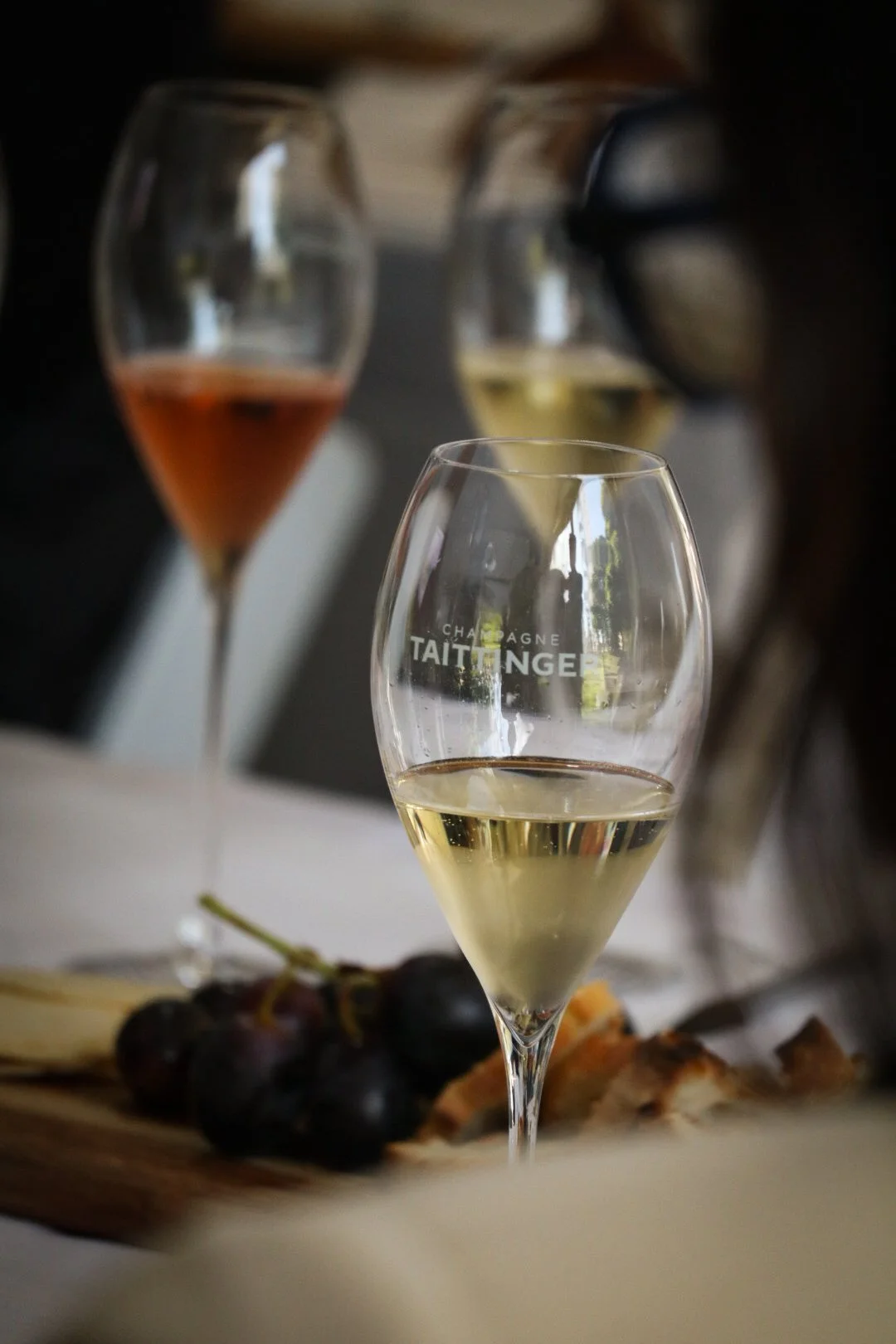 Verres de champagne blanc et rosé avec des raisins noirs et du fromage sur une planche, floutés en arrière-plan.