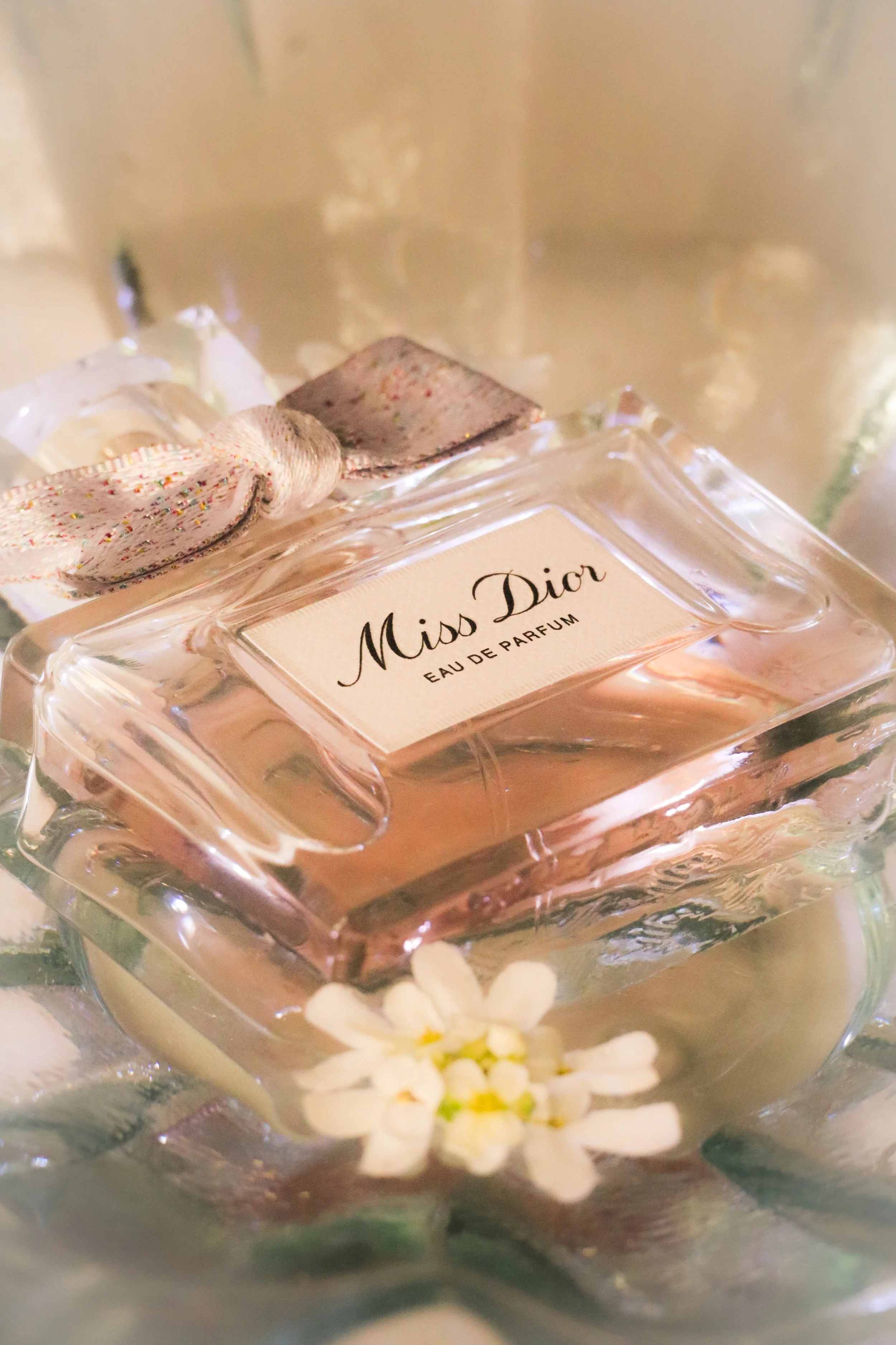 Une bouteille de parfum Miss Dior dans un flacon en verre avec un ruban multicolore noué autour du cou, avec des petites fleurs blanches en premier plan.
